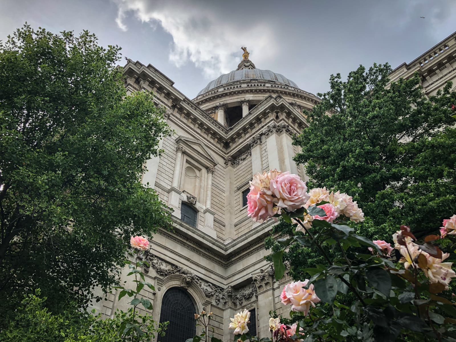 An upward view of a grand architectural building, likely a cathedral, partially obscured by lush green trees and blooming pink roses in the foreground. The ornate dome of the building is topped with a golden figure, standing out against a cloudy sky, while the detailed stonework of the structure adds an element of historical grandeur.