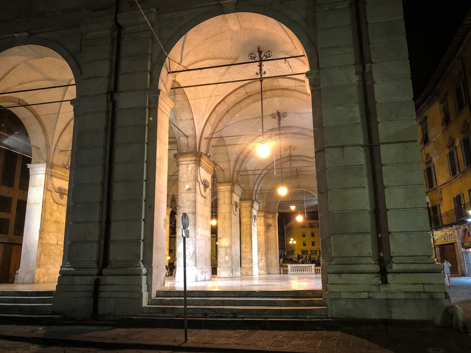 A nighttime view of a grand stone archway, illuminated by soft yellow lights. The tall, imposing columns support an arched ceiling, with hanging lights adding a warm glow. The surrounding area is dimly lit, with a glimpse of nearby buildings and people in the distance under the archway.