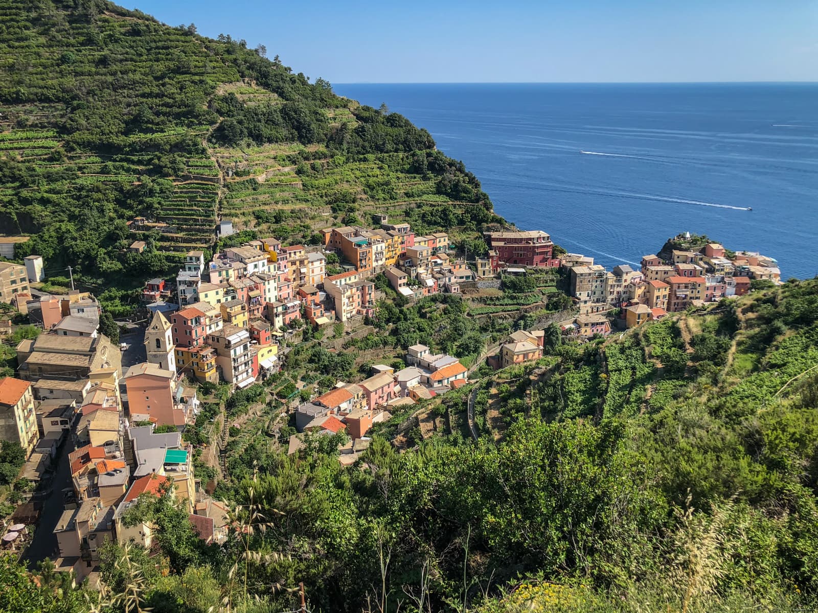A bird's-eye view of a cliffside village with tightly clustered colorful houses. The village nestles along the steep hillside, surrounded by terraced fields and overlooking the calm, azure ocean.