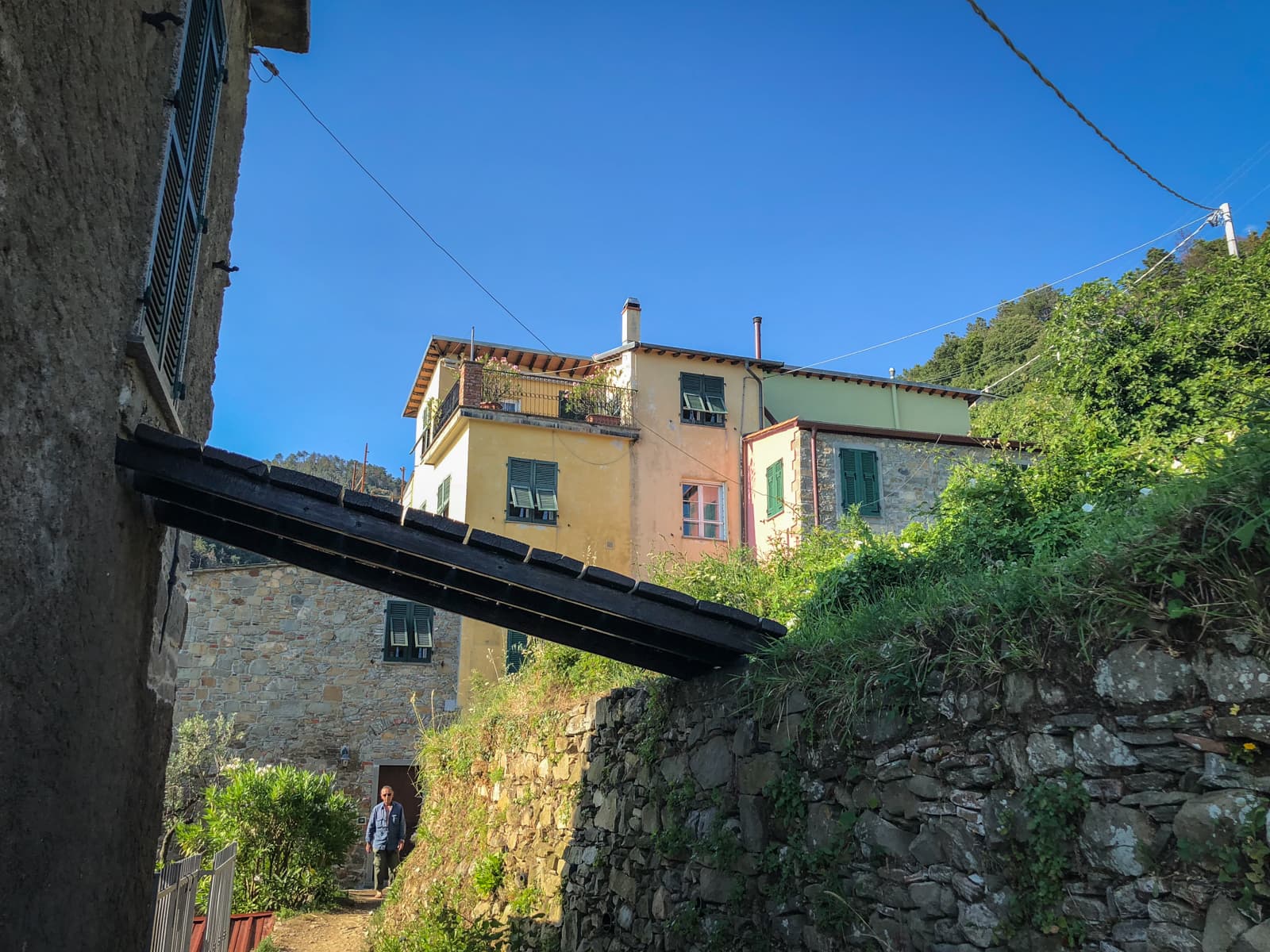 A peaceful village scene, where colorful houses sit on a hillside under a clear blue sky. A person is seen walking along a path, adding a sense of quiet and everyday life.