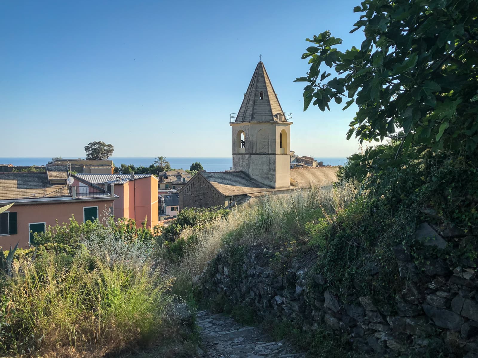 A charming church tower in a small village with the blue sea visible in the background. The image has a calm, picturesque feeling, with rustic houses lining the narrow pathways of the village.