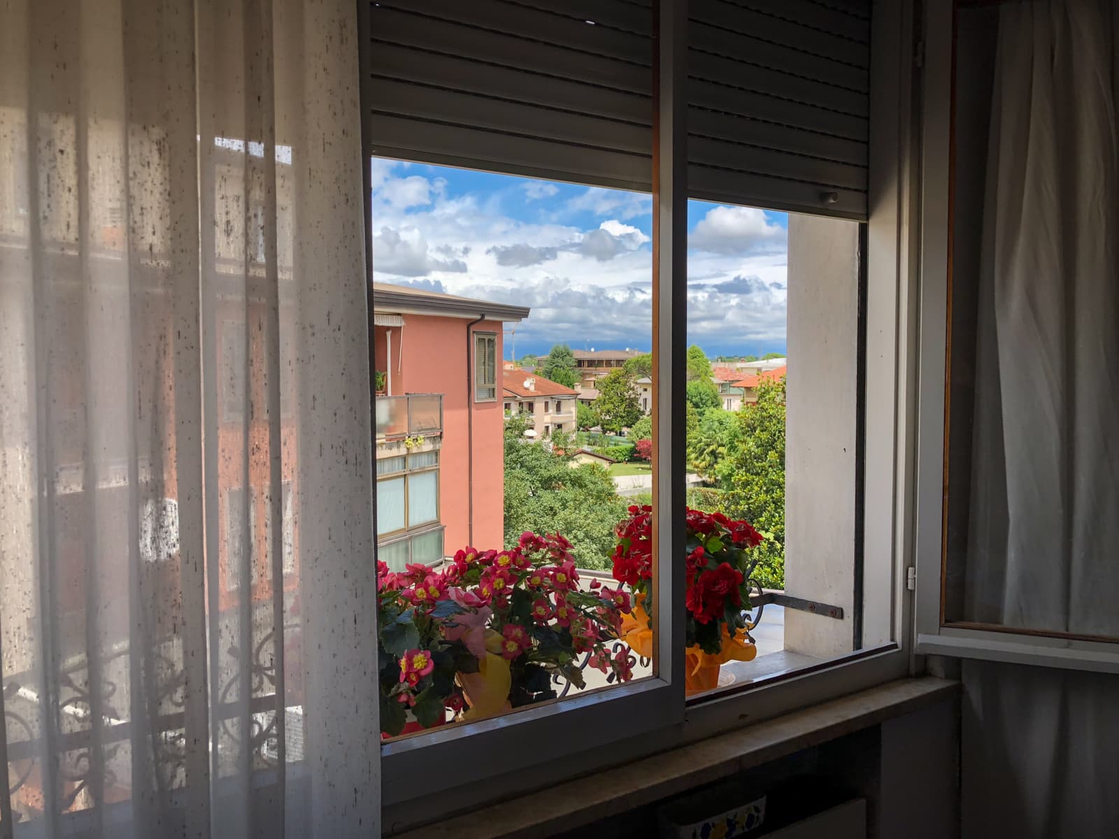 The image shows a view from a partially open window with light curtains on one side. Outside, there is a vibrant display of red and pink flowers in pots on the windowsill. In the background, you can see a neighborhood with several buildings, trees, and greenery. The sky is partly cloudy, with patches of blue and fluffy white clouds, adding a peaceful atmosphere to the scene.