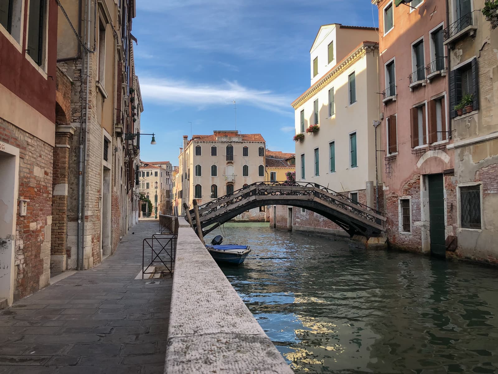 A quiet and charming Venetian canal with a small arched pedestrian bridge crossing over the water. The narrow canal is lined with historic buildings, their windows adorned with flower pots. A boat is moored by the side, and the scene is bathed in soft sunlight under a clear blue sky.