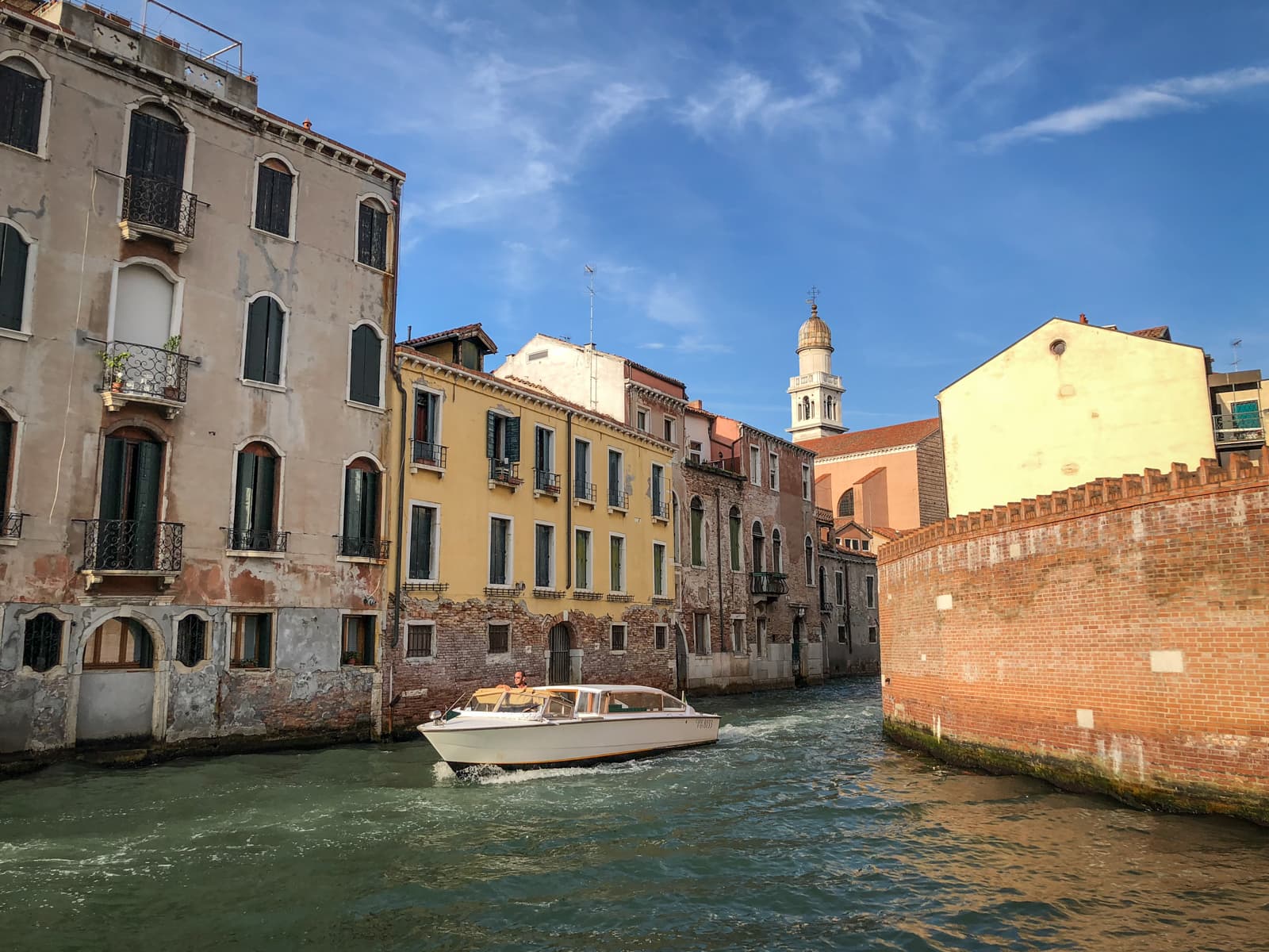 A vibrant canal scene in Venice, with a motorboat cruising along the greenish water. On both sides of the canal are old, weathered buildings, some with faded facades and others painted yellow. Small balconies and shutters add character to the buildings. In the background, a church tower rises against a clear blue sky.