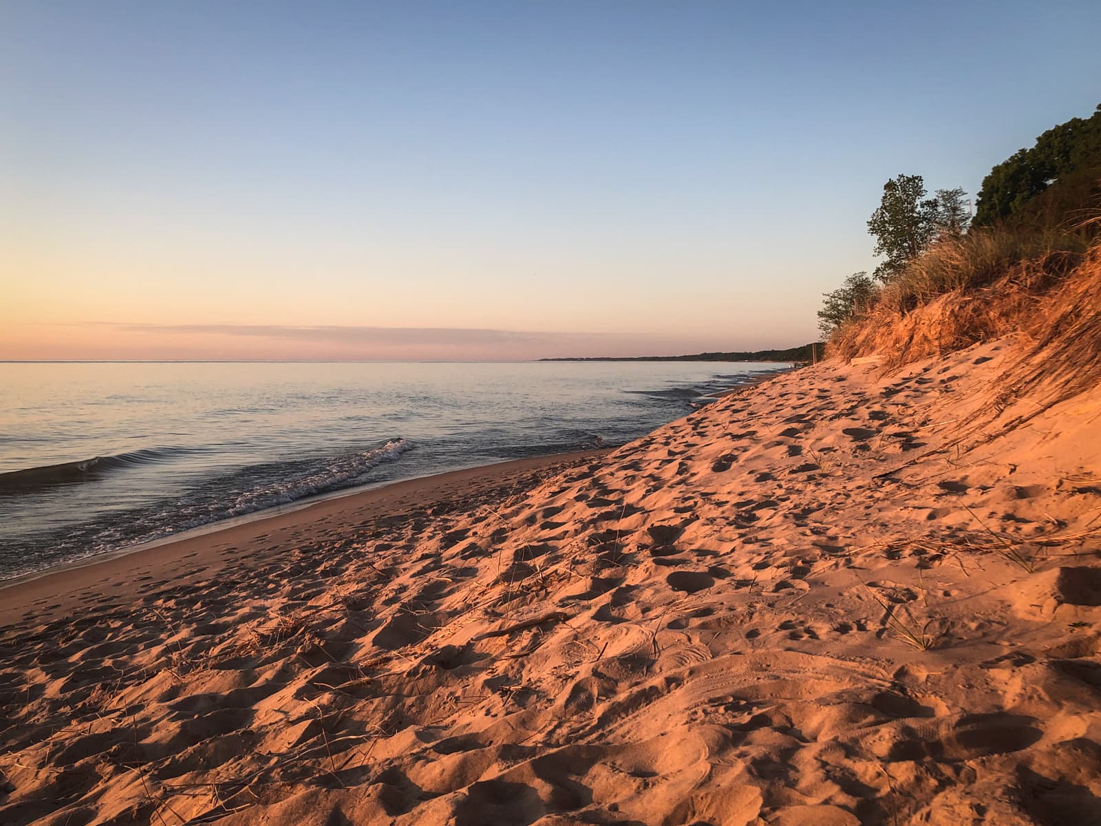 A peaceful beach at sunset, with soft waves gently lapping at the shore. The sandy beach is bathed in a warm golden light, with footprints scattered across the sand. Grassy dunes rise on the right side of the image, while the calm water stretches out to the horizon. The sky is clear, with soft pastel hues of orange and pink fading into the blue as the sun sets, creating a serene and tranquil atmosphere.