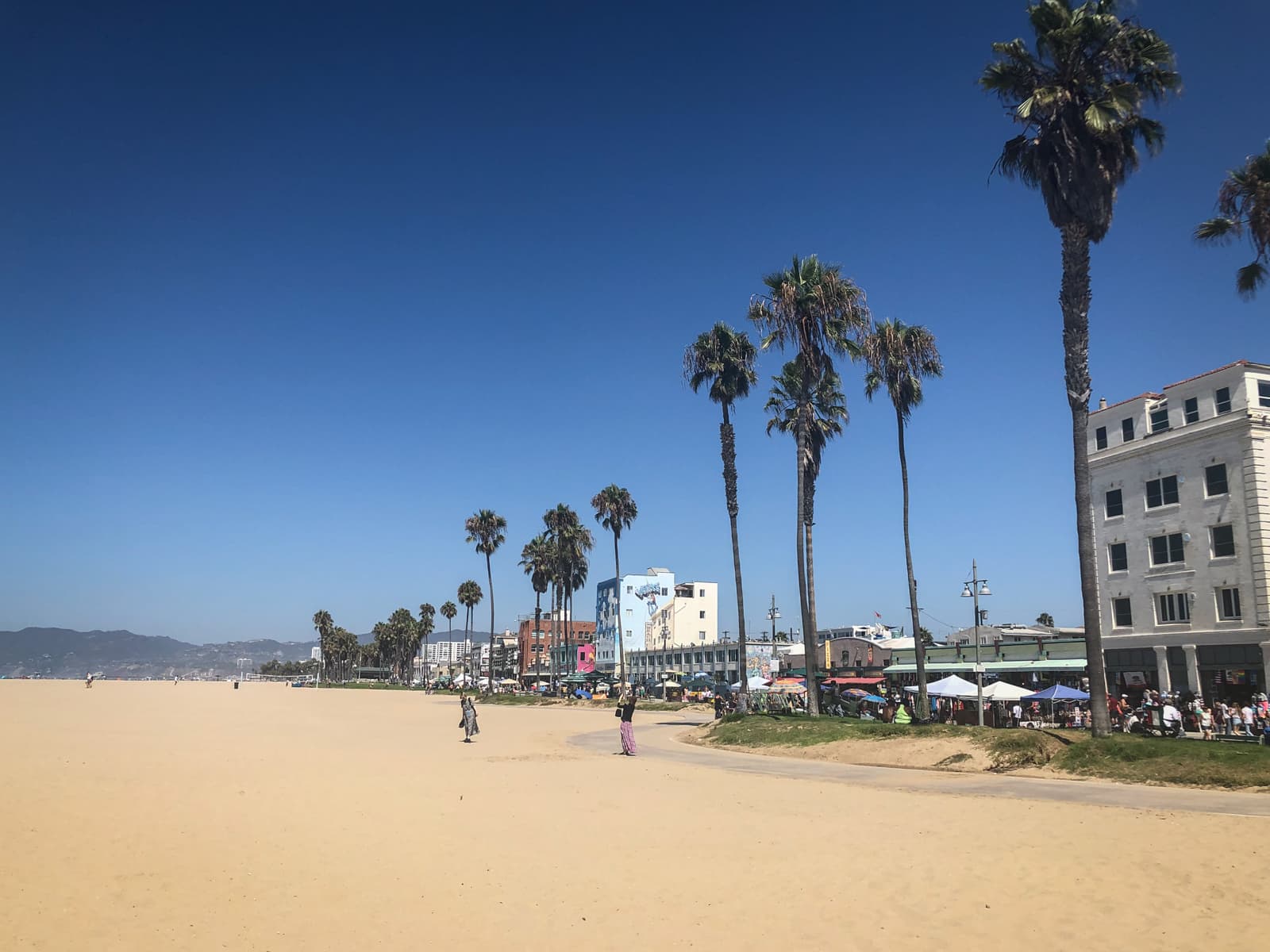 A sunny beach scene with a wide stretch of golden sand under a clear blue sky. Tall palm trees line a walkway on the right side, leading to a lively area with shops, cafes, and people enjoying the day. The buildings along the beachfront have a mix of bright colors and classic architecture, with mountains visible in the distance. The beach is sparsely populated, with a few people walking along the sand, creating a laid-back, vibrant atmosphere.