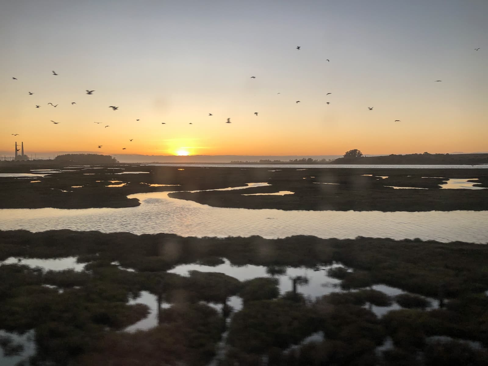 A tranquil sunset over a marshland with patches of water reflecting the soft light. The sky is a gradient of orange and yellow as the sun dips below the horizon. A flock of birds is in flight across the sky, adding movement to the peaceful scene. In the distance, silhouettes of trees and industrial structures, including a pair of tall chimneys, can be seen, contrasting with the natural landscape. The calm waters and open space evoke a serene and quiet atmosphere.