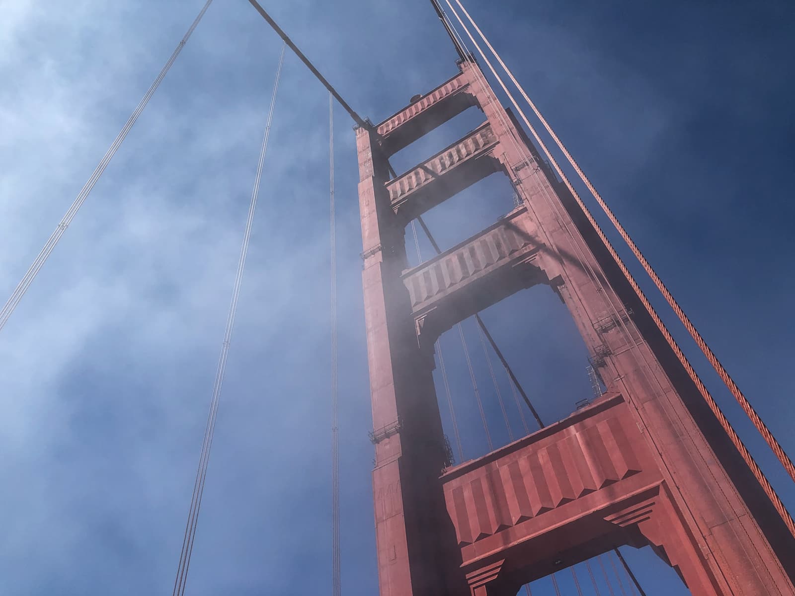 A view looking up at one of the tall towers of a red suspension bridge. The thick cables run along the tower, which stands against a backdrop of blue sky and soft clouds or mist. The sunlight casts shadows, adding depth to the architectural features of the tower.