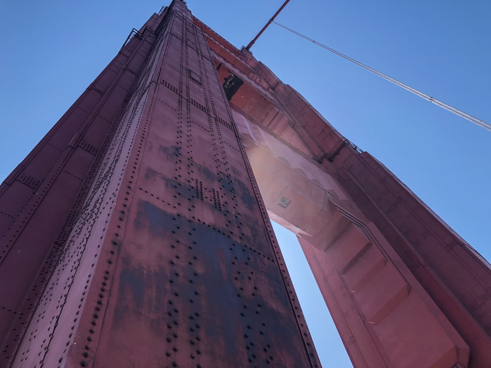 A close-up, upward view of a massive red tower of a suspension bridge. The riveted steel beams and weathered surface of the tower are bathed in sunlight, with cables extending from the structure into the clear blue sky above. The sheer size and industrial details of the tower dominate the image.