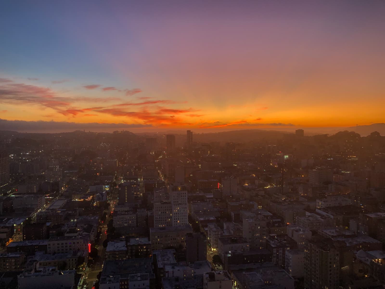 A breathtaking cityscape at sunset, with the sky awash in vibrant shades of orange, pink, and purple as the sun dips below the horizon. The city below is beginning to light up for the evening, with illuminated streets and buildings visible as the twilight deepens. In the distance, hills and a faint outline of water are visible, with the soft glow of the sunset casting a warm hue over the entire scene.