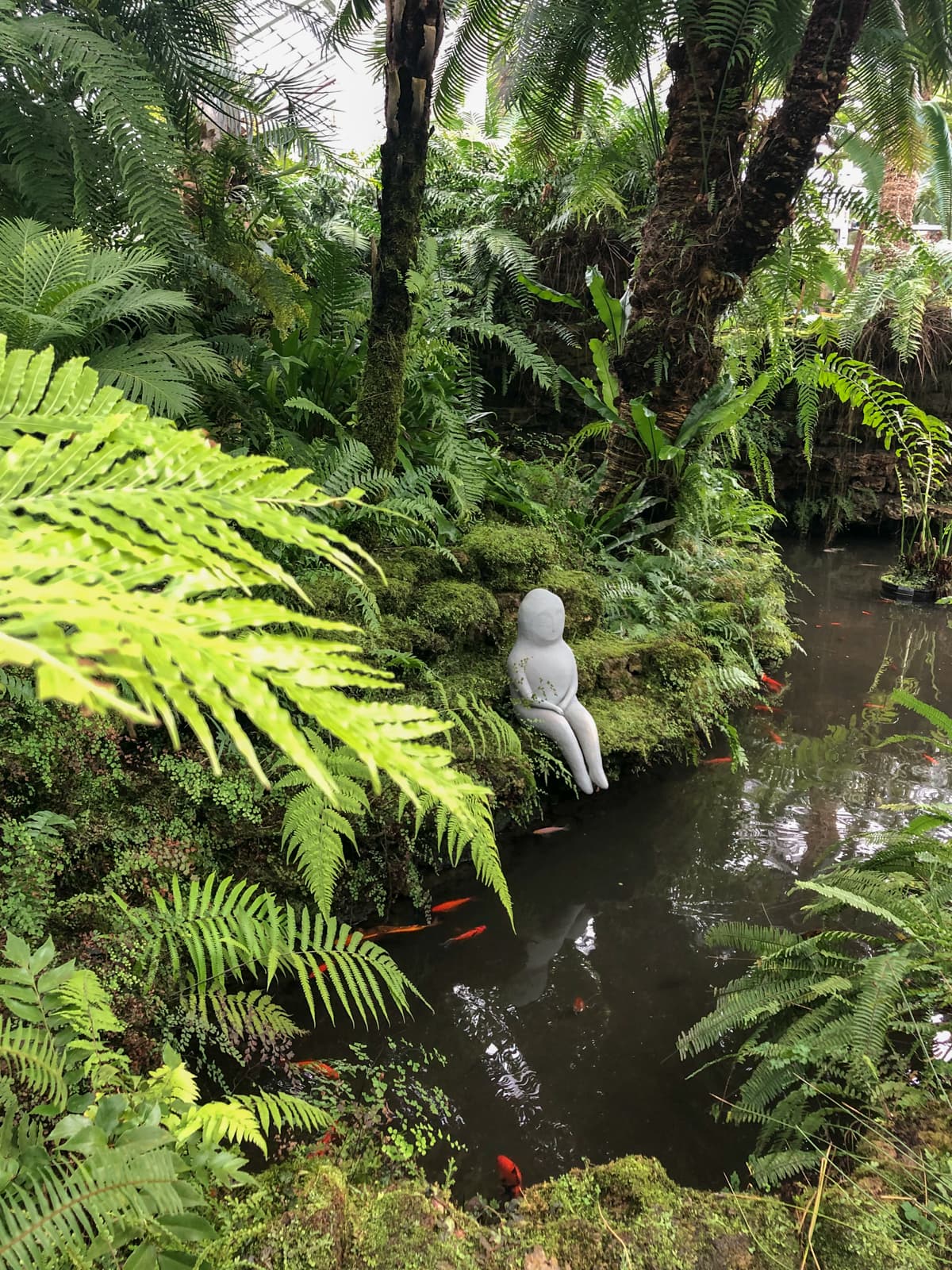 A lush, tropical scene featuring a small pond surrounded by dense greenery, including ferns, moss, and palm trees. In the center of the image, a serene white sculpture of a seated figure rests on a moss-covered rock by the water’s edge. Bright orange fish swim in the calm pond, adding a splash of color to the tranquil setting. The vibrant plants and peaceful atmosphere create a secluded, meditative garden scene.