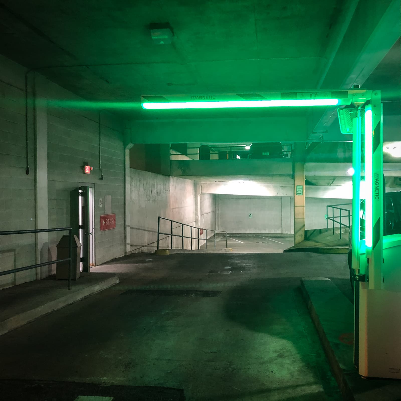 An empty parking garage with a dimly lit, industrial atmosphere, illuminated by a bright green fluorescent light hanging from the ceiling. The green light casts an eerie glow over the concrete walls and floors. In the foreground, a barrier arm is partially visible, and a small side door is open to the left. The muted colors and shadows create a stark, almost unsettling feeling within the enclosed space.