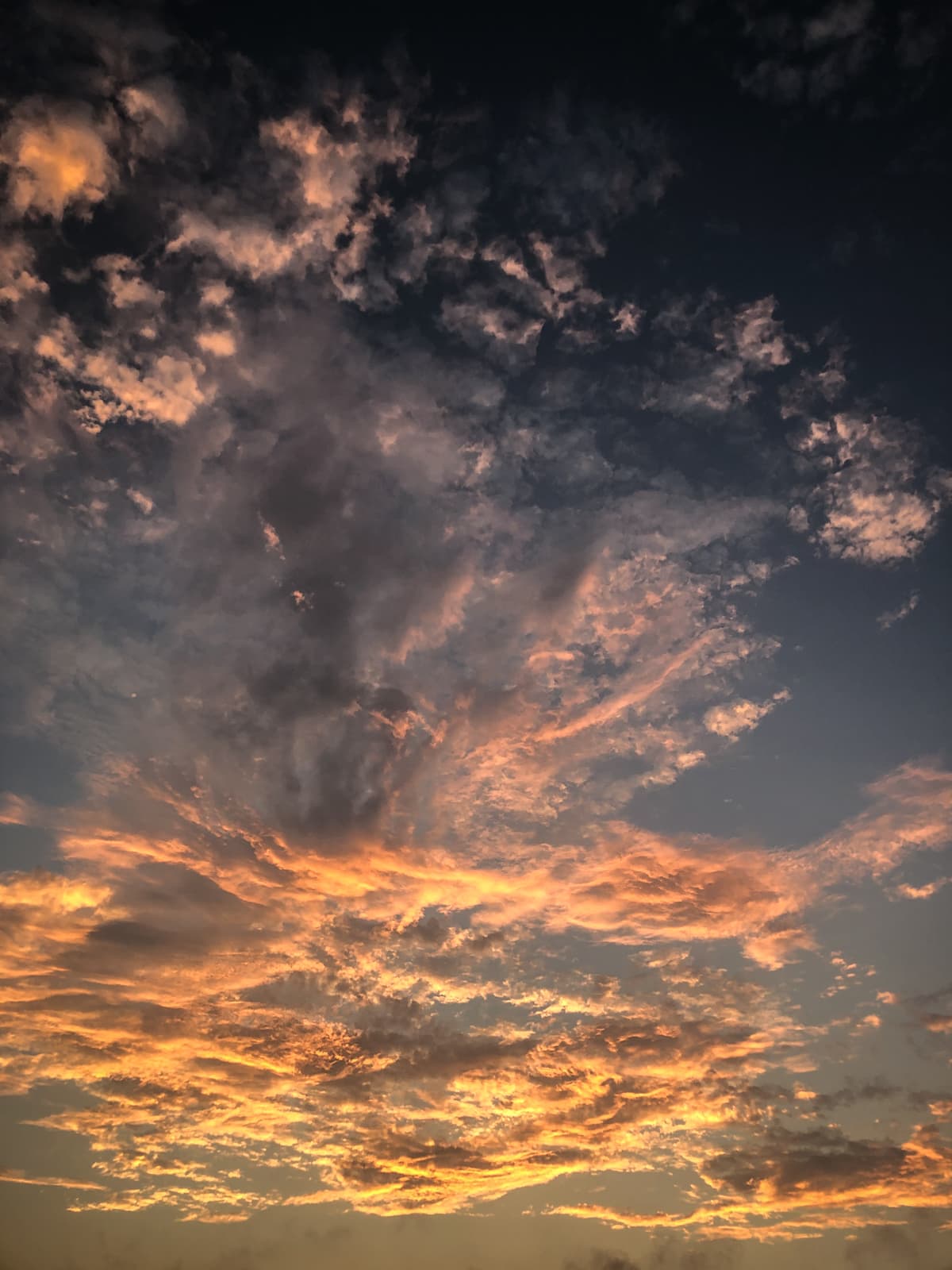 A stunning sunset sky filled with dramatic clouds illuminated by golden and orange hues. The clouds are spread across the sky in various formations, with the light from the setting sun casting a warm glow on the lower portion of the clouds, creating a beautiful contrast against the darker, shadowed clouds higher up. The scene captures the peaceful and awe-inspiring beauty of a sunset.