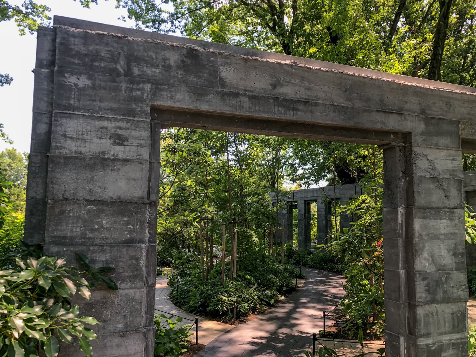 A shaded garden pathway bordered by tall plants and trees, winding through a series of concrete archways. The sunlight filters through the dense foliage, casting soft shadows on the path. The weathered concrete structures add a contrasting modern, architectural element to the natural surroundings.
