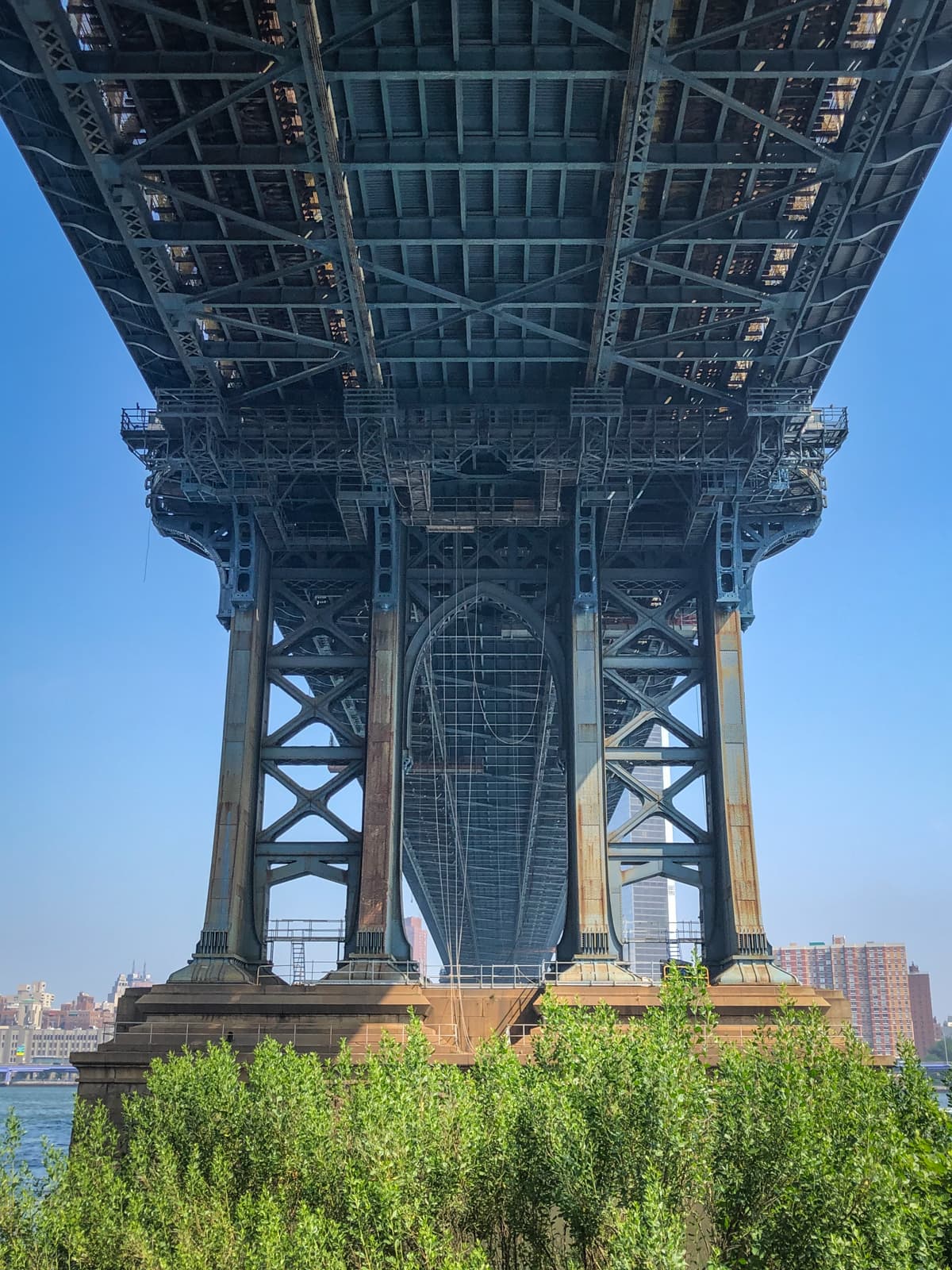 A dramatic view from beneath a large suspension bridge, showcasing the intricate metal framework and beams that support the structure. The massive pillars of the bridge rise from a base surrounded by greenery, with the city skyline faintly visible in the background under a clear blue sky. The perspective emphasizes the scale and engineering of the bridge, creating a sense of both strength and complexity.