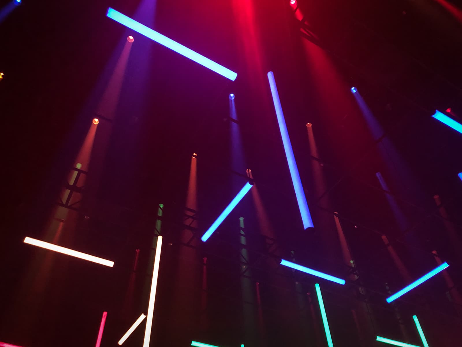 An abstract and vibrant display of colorful neon lights suspended from the ceiling in a dark space. The lights are arranged in various directions, with hues of blue, red, green, and pink glowing brightly against the shadows. The beams of light create a dynamic and futuristic atmosphere, with each color casting a subtle glow onto the surrounding area. The overall effect is energetic and visually striking, resembling a modern light installation or art piece.