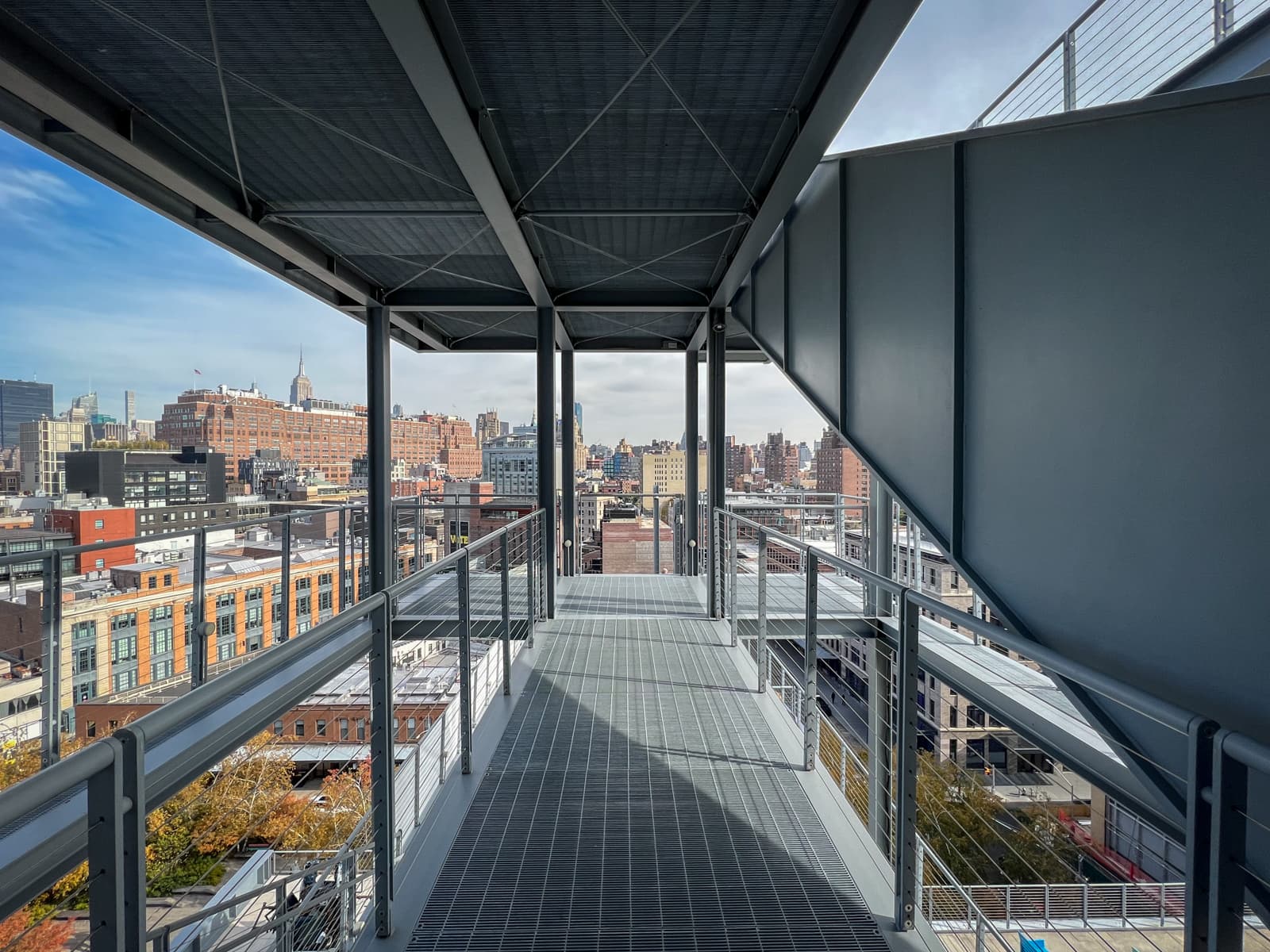 A metal walkway with railings leads to an open view of the city skyline. Modern buildings and rooftops are visible in the distance, with the Empire State Building standing tall in the background against a clear sky.