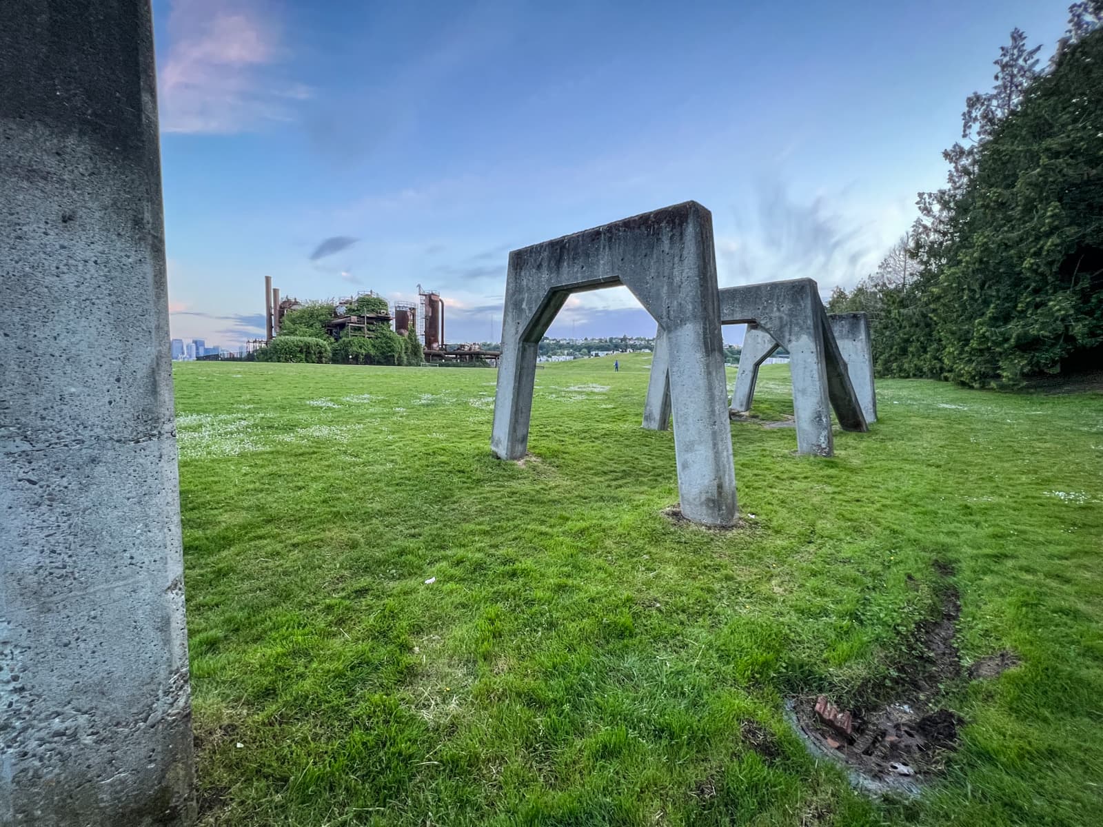 A wide shot of a grassy field dotted with large, industrial concrete structures that resemble arches or frames. In the background, there is an overgrown industrial plant, partially hidden by greenery, with a few distant buildings visible under a soft, pastel-colored sky.