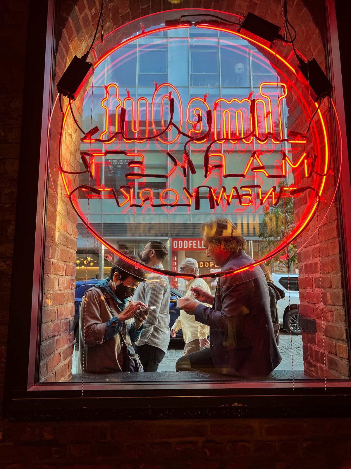 A vibrant image of people sitting outside a restaurant, seen through a large window framed by exposed brick. The neon red "Time Out Market New York" sign is glowing, with the text reversed from the outside view. The street outside is shown through the glass, showing passersby and nearby storefronts. The warm glow of the neon sign contrasts with the cooler tones of the exterior, creating a lively, urban atmosphere that blends both the indoor and outdoor worlds.
