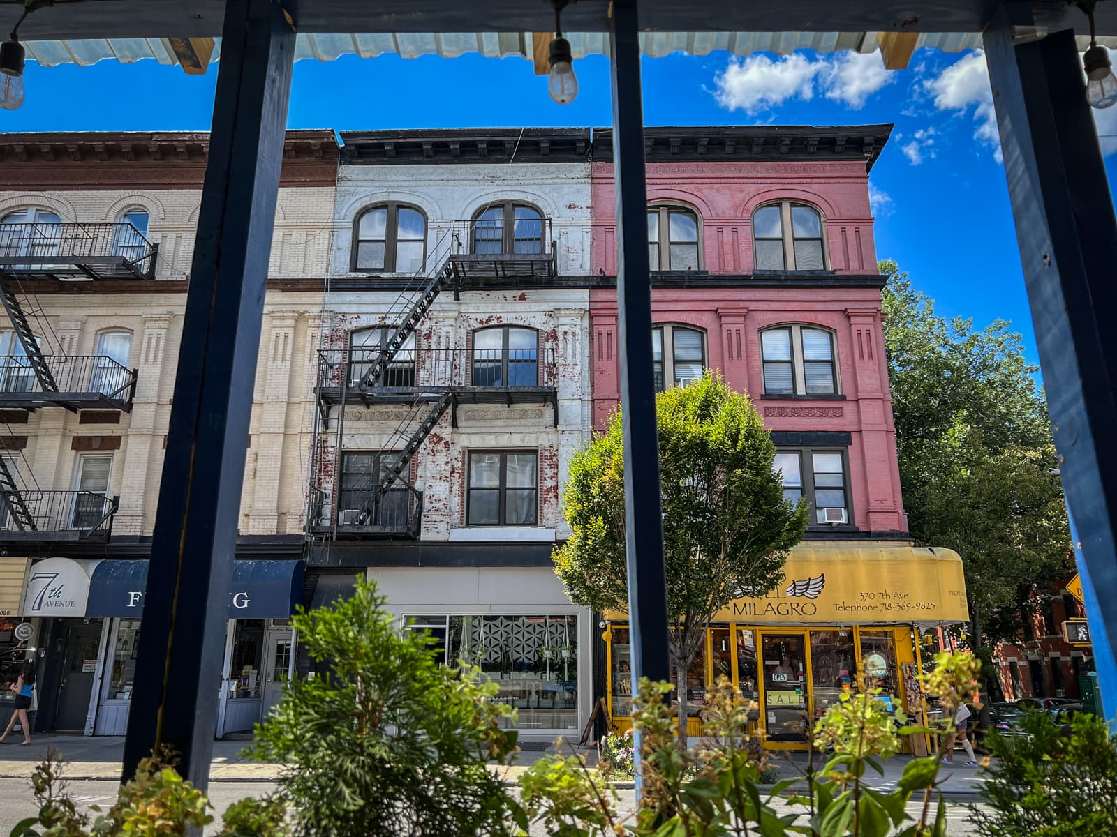 A sunny street scene featuring a row of colorful, old-fashioned buildings with fire escapes. The building on the right is painted pink with black accents, while the middle building has a weathered white facade with visible brick. The storefronts below include a bright yellow shop called "Milagro" and a blue awning over a shop labeled "7th Avenue." Green plants in the foreground frame the scene, adding a natural contrast to the urban architecture. The vibrant blue sky with scattered clouds provides a cheerful backdrop to the lively neighborhood.