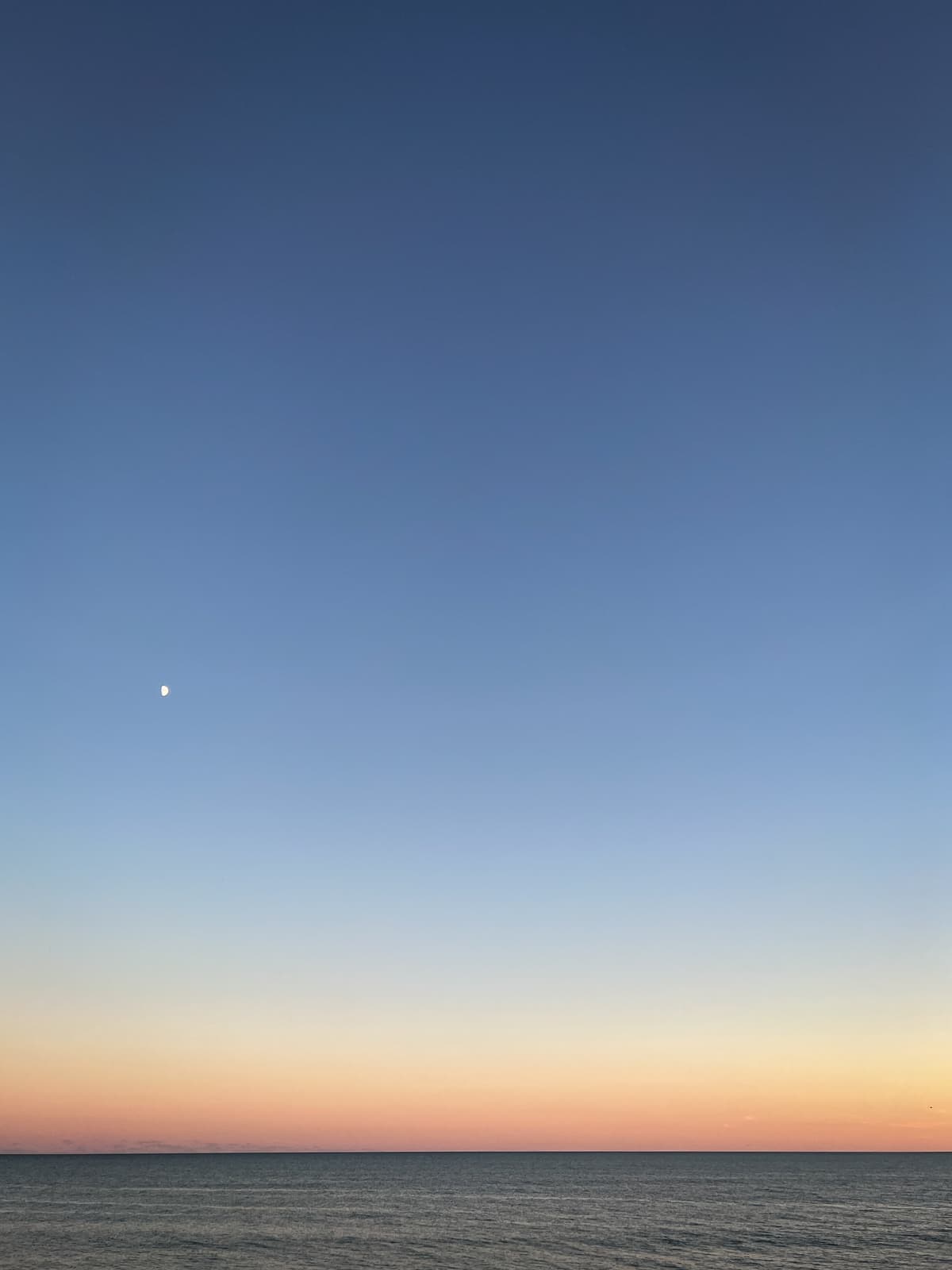 A serene image of a vast ocean horizon at sunset, with the sky transitioning from soft pastel orange near the horizon to deep blue higher up. A small, bright moon is visible in the upper left corner, contrasting with the fading daylight. The calm water reflects the warm hues of the setting sun, creating a peaceful and minimalist scene, emphasizing the tranquility of nature.