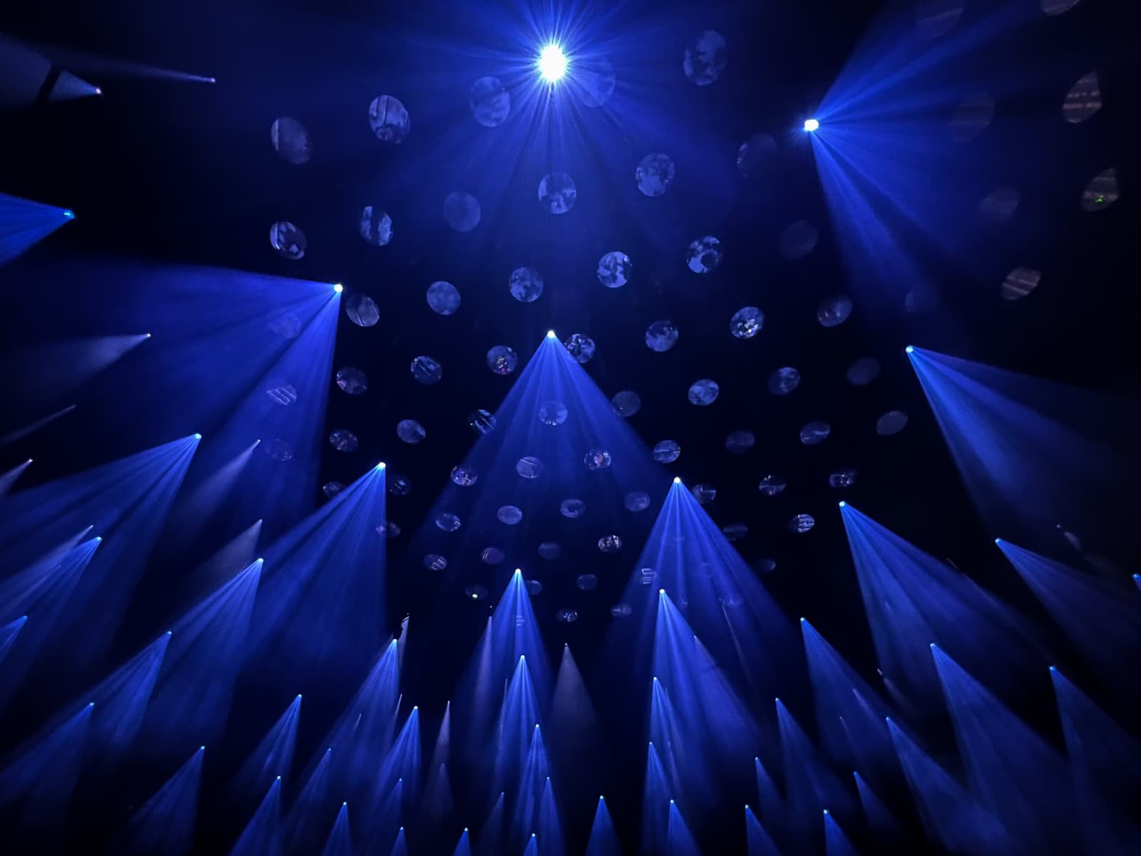 A dramatic light show with beams of blue light extending from above, creating a radial pattern. The lights shine down onto a darkened space, highlighting circular reflective surfaces suspended in the air. The vivid blue lighting and the ethereal atmosphere evoke a futuristic, immersive experience.