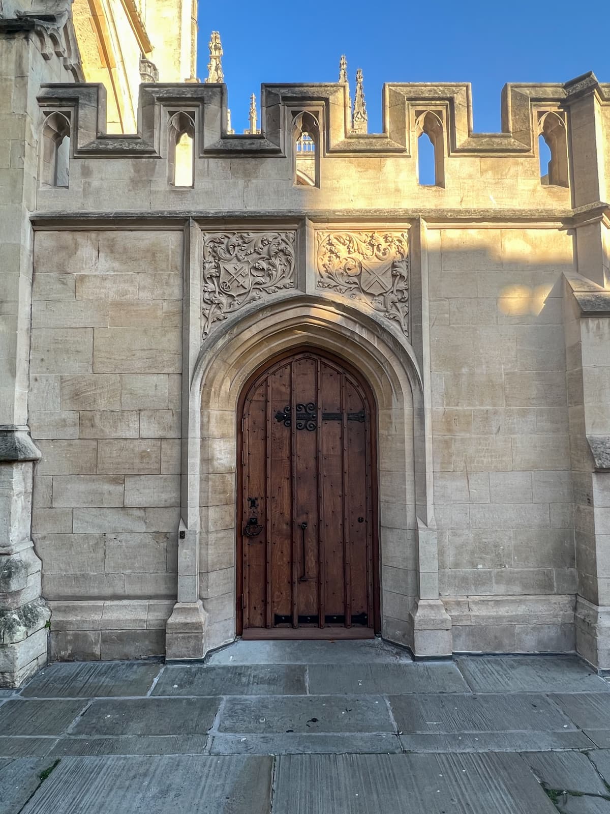 A close-up view of an ornate stone doorway on a historic building. The arched wooden door is framed by detailed stone carvings, including crests and floral designs. Above the door, a row of battlements with narrow windows adds a medieval feel to the architecture. The sunlight casts soft shadows on the light-colored stone, highlighting the craftsmanship and elegance of the structure.