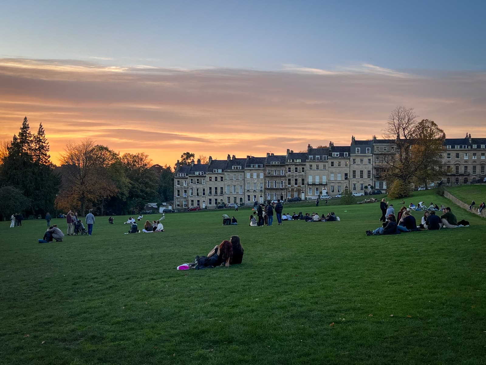 A tranquil sunset scene in a park, with groups of people relaxing on the expansive grassy field. In the background, historic buildings with stone facades line the horizon, and the sky is filled with warm orange and pink hues, transitioning into a soft blue. The trees in the park add an autumnal touch, with some displaying golden leaves as the sun sets.
