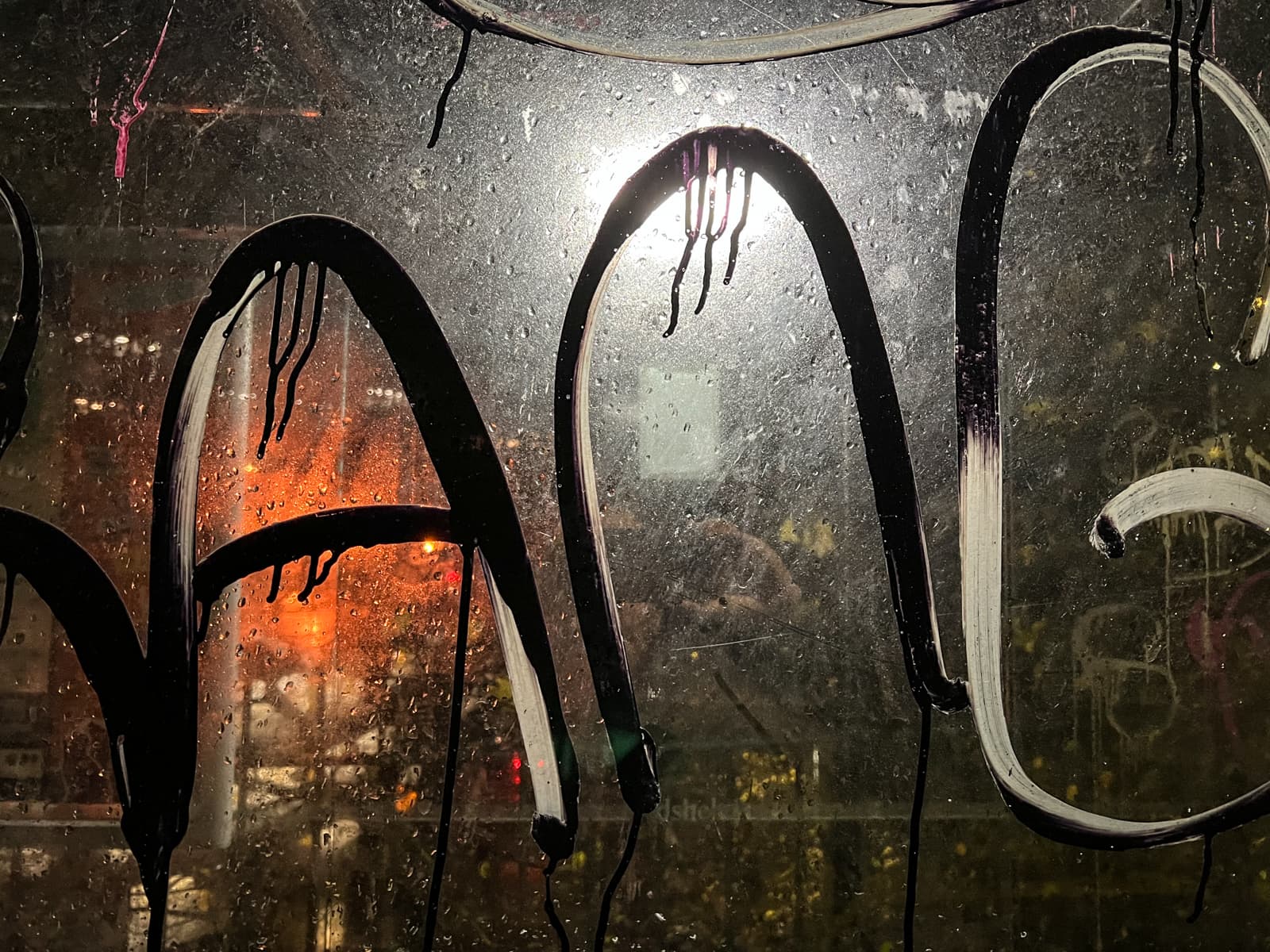 A close-up of a glass window covered in large black graffiti, with light shining through from behind, highlighting the droplets of moisture and creating a wet, gritty texture. An orange light glows faintly in the background.