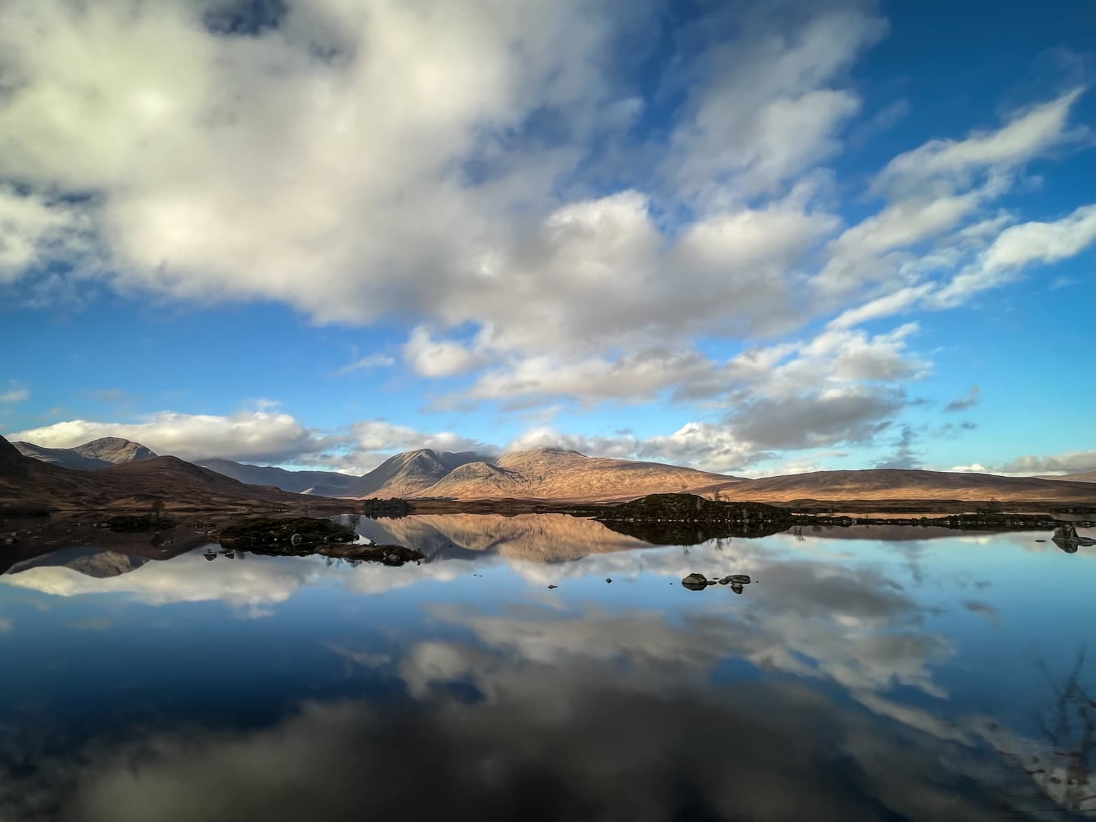 A tranquil lake reflecting the mountains and clouds perfectly on its glassy surface. The surrounding hills are bathed in soft sunlight, and the sky is a brilliant blue, dotted with fluffy clouds.