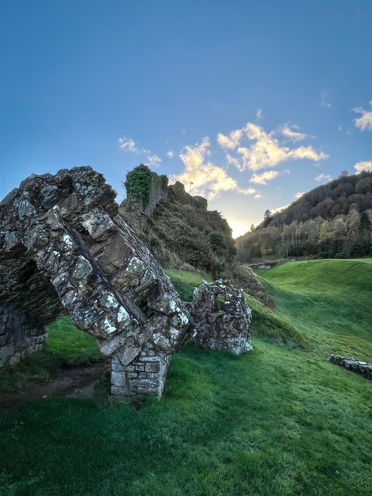 A fragment of an ancient stone archway set on a grassy hill. The structure appears to have partially collapsed, and beyond it, more stone ruins and green hills can be seen under a clear sky.