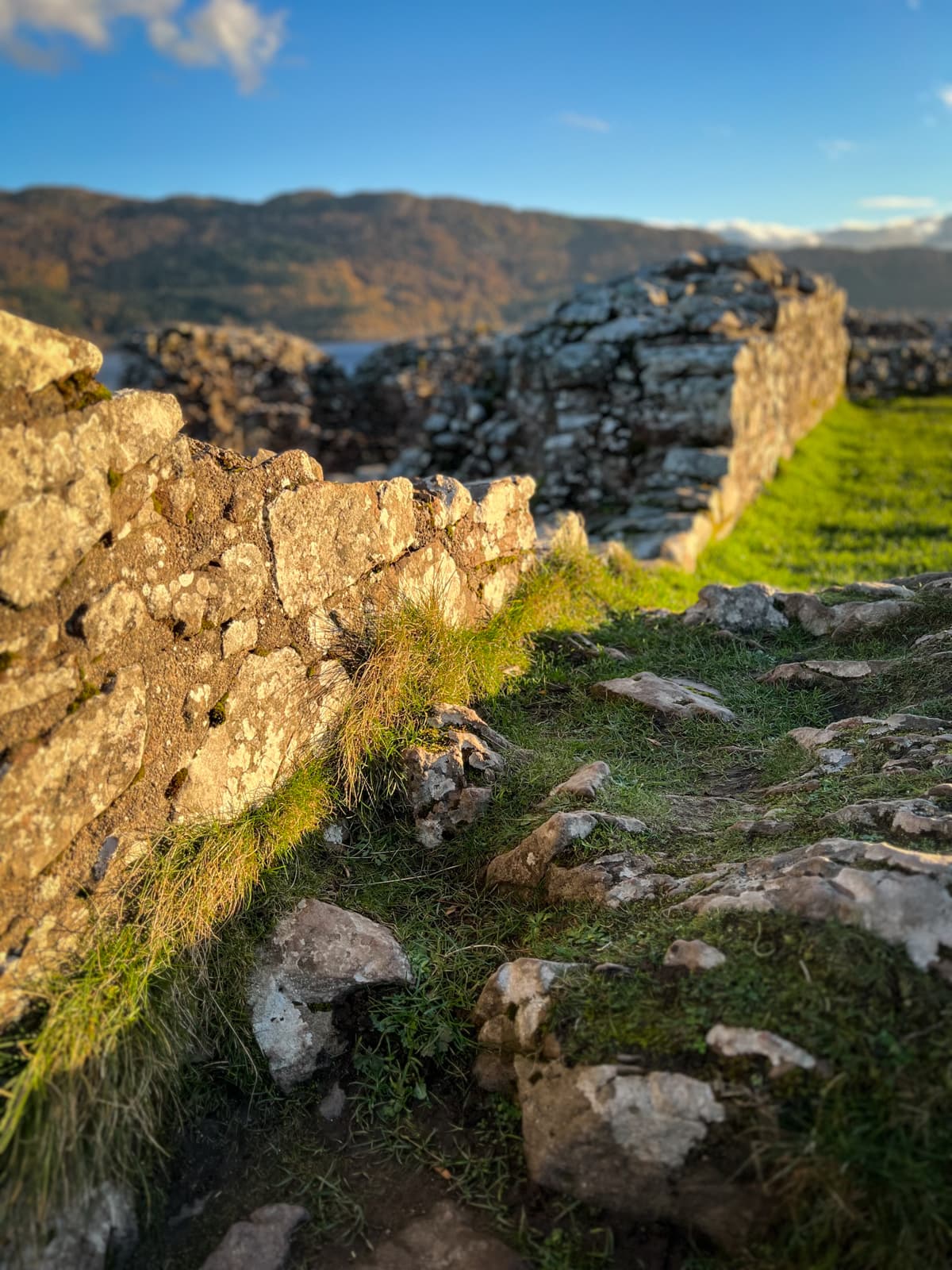 A close-up of crumbling stone walls covered with patches of grass and moss. The weathered stones appear to form part of an ancient structure, with distant hills visible in the background.