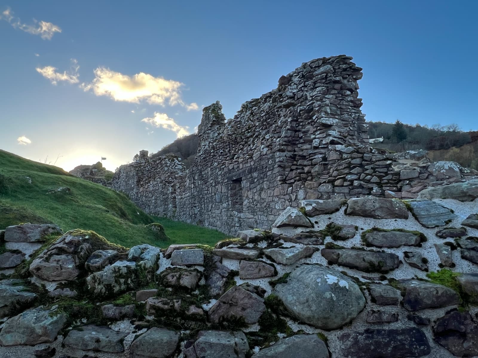 A stone ruin standing on a green grassy hillside, with the sun setting behind the structure. The silhouette of the stone walls stands out against the golden light filtering through the clouds.