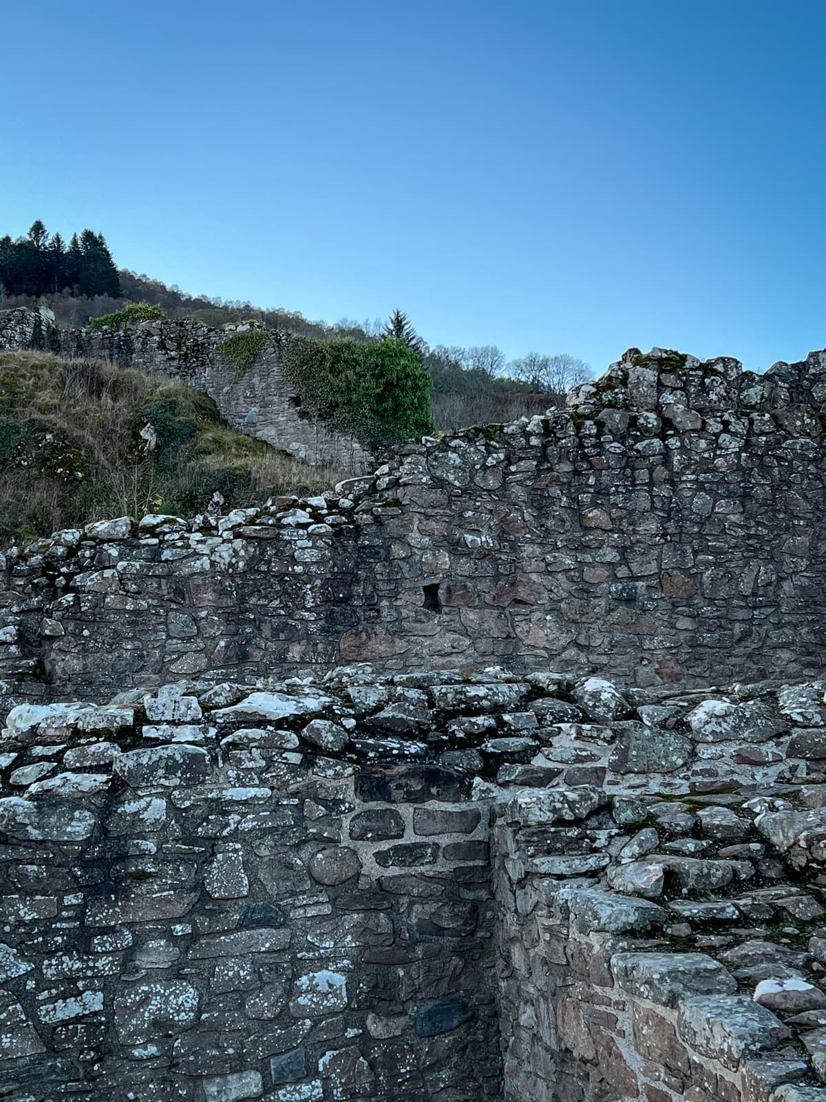 Ancient stone ruins standing against a bright blue sky. The walls are weathered and overgrown with some greenery, with a backdrop of trees and hills.