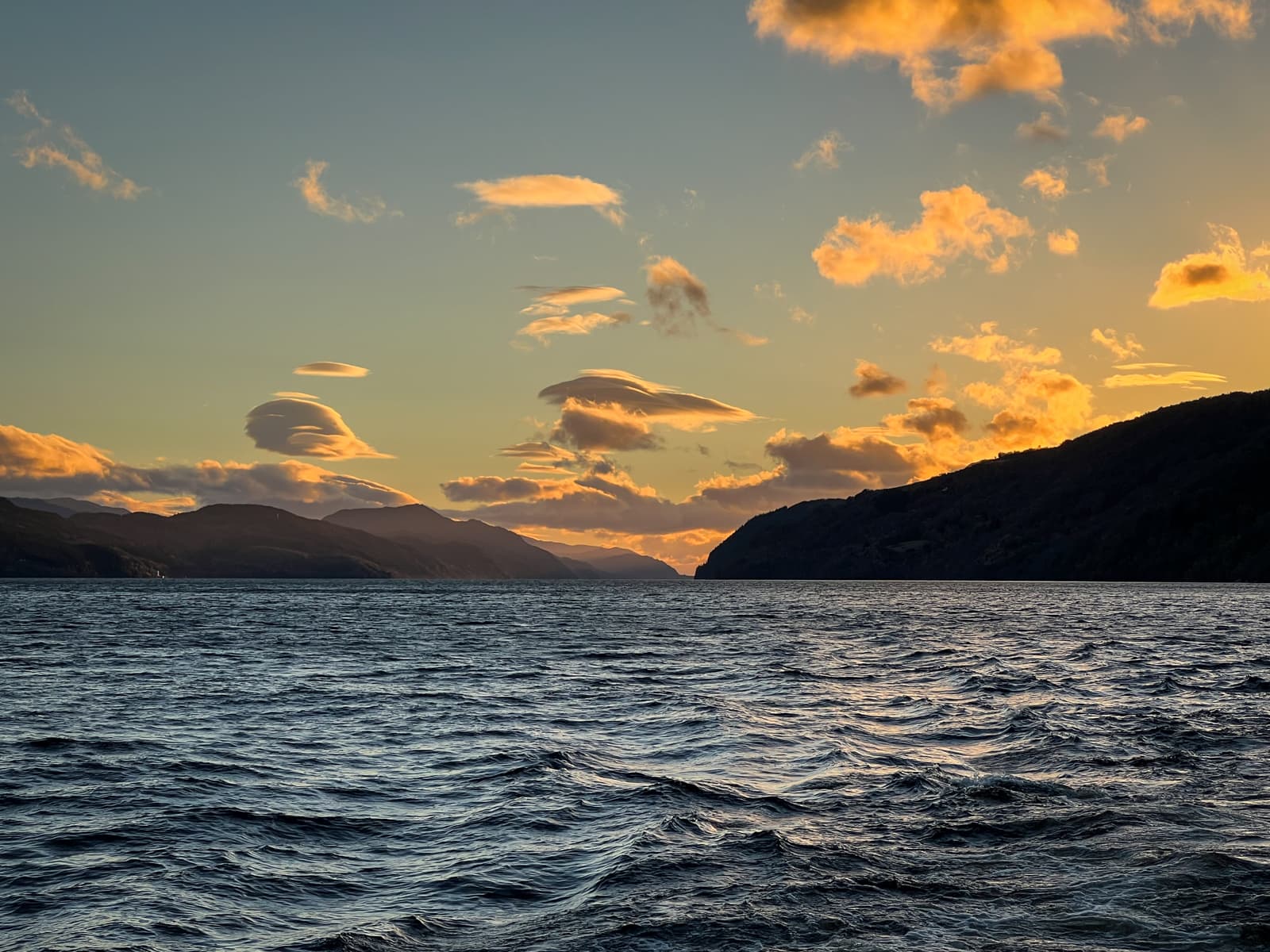 A serene view of a large body of water surrounded by hills at sunset. The sky is filled with scattered clouds illuminated by the warm glow of the setting sun, casting soft reflections on the water's surface.