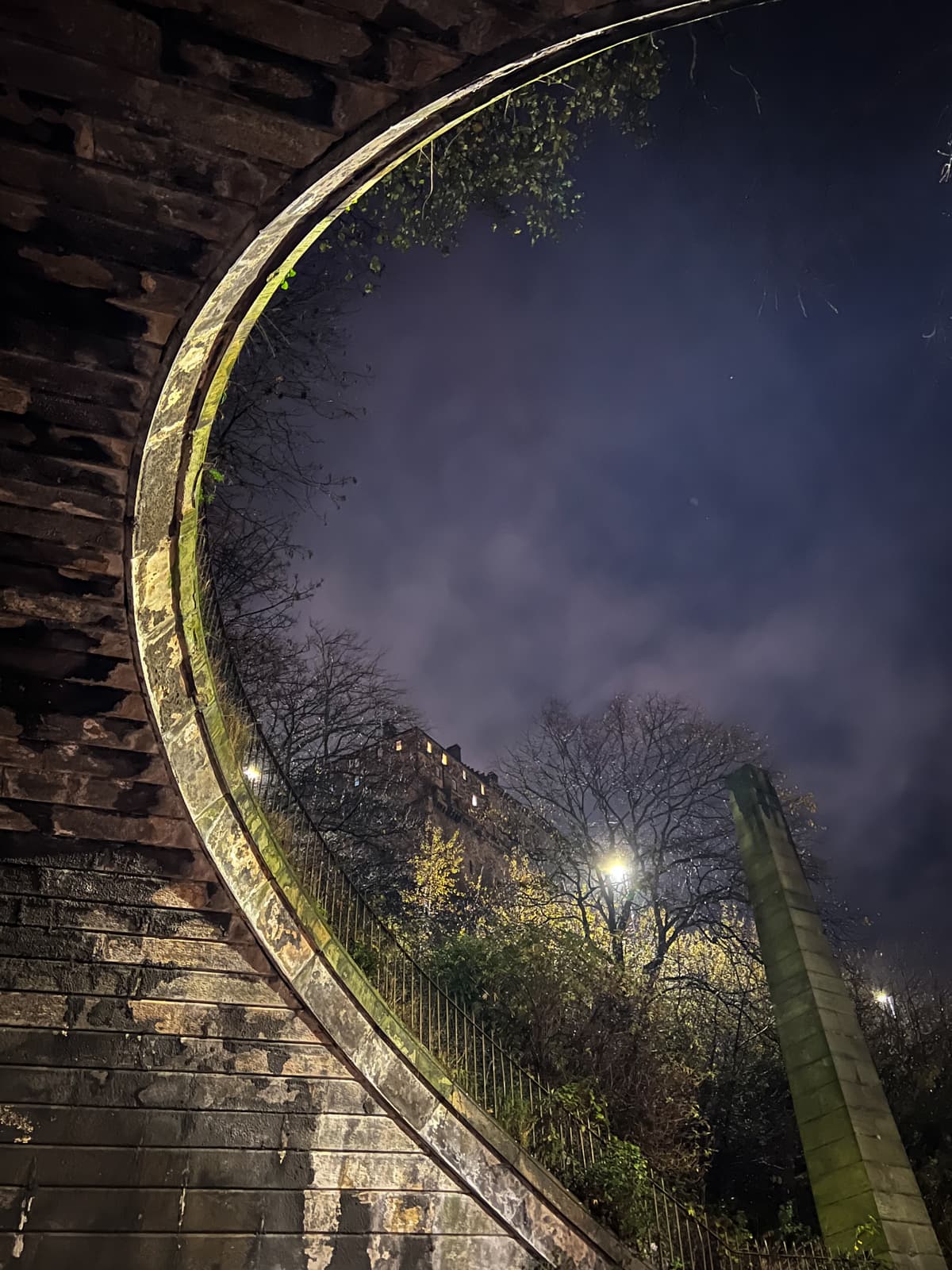 A night view looking up through an archway. The stone arch frames a dark, cloudy sky, and tree branches are faintly visible, illuminated by dim streetlights. A stone column stands near the edge of the frame, partially lit.