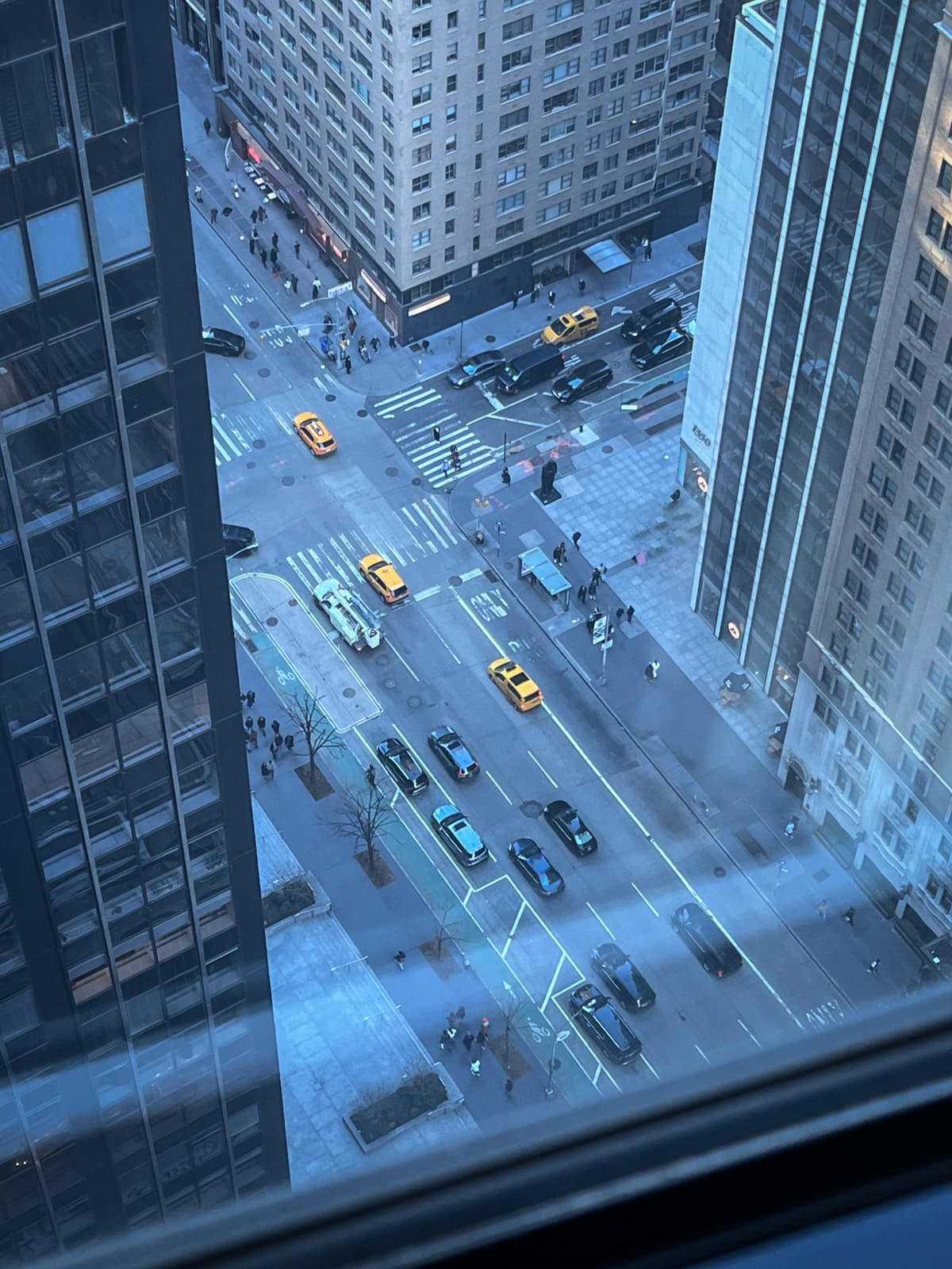 A bird's-eye view of a busy city street intersection, with several yellow taxis and cars driving along the road. Pedestrians can be seen walking along the sidewalks, and the scene is framed by tall buildings, including glass-covered skyscrapers. The streets feature marked bike lanes and crosswalks, with traffic flowing smoothly. The cool tones of the photo, possibly taken during the early morning or late afternoon, create a calm urban atmosphere.
