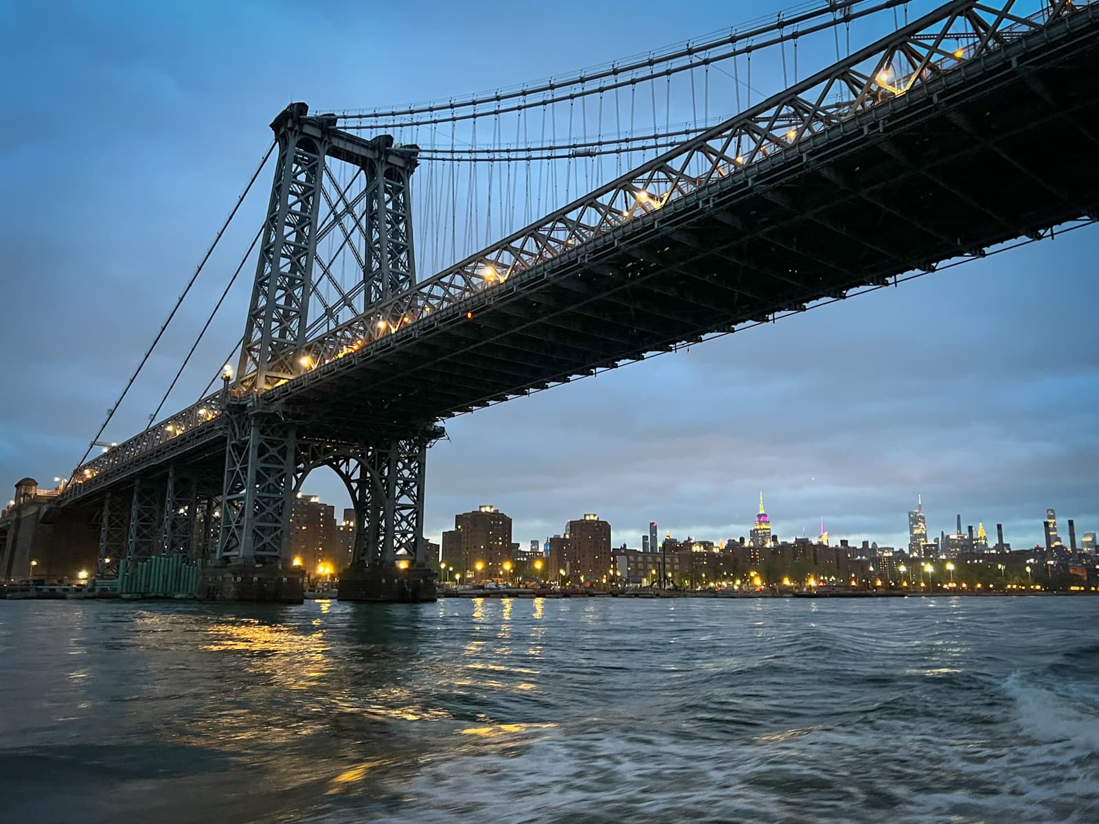 A photo of a large suspension bridge at dusk, its structure illuminated by lights. The skyline of a city with glowing buildings is visible in the distance across the water, and the surface of the water reflects the city lights.