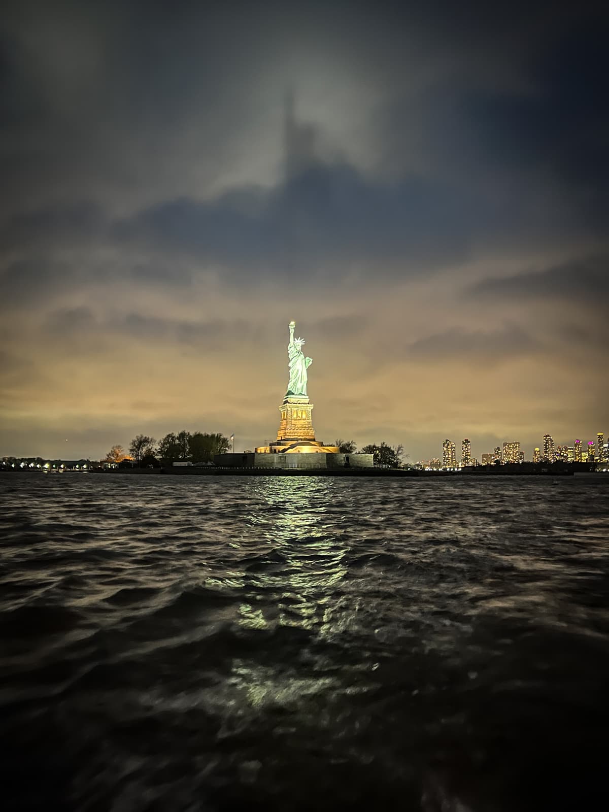 A nighttime photo of the Statue of Liberty illuminated against a cloudy sky. The statue stands tall on Liberty Island, with the dark waters of the harbor in the foreground reflecting the statue's light. In the distant background, the lights of the city skyline are faintly visible.