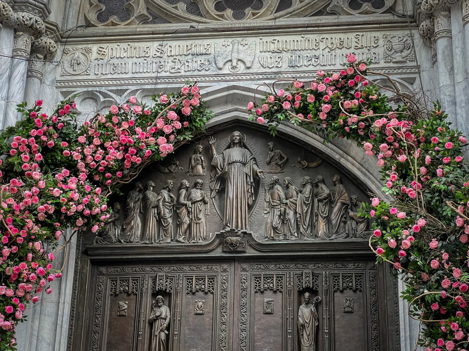 This image shows an ornate religious or historical doorway, decorated with a beautiful arrangement of pink roses, forming an arch around the entrance. The doorway itself features intricate stone carvings and relief sculptures of figures, possibly saints or apostles, arranged around a central figure, likely representing Christ. Above the door, there is Latin text engraved, which translates to spiritual or religious phrases. The arch and the floral decorations enhance the solemnity and beauty of the architectural design, adding a vibrant touch of nature to the otherwise stone-heavy structure.