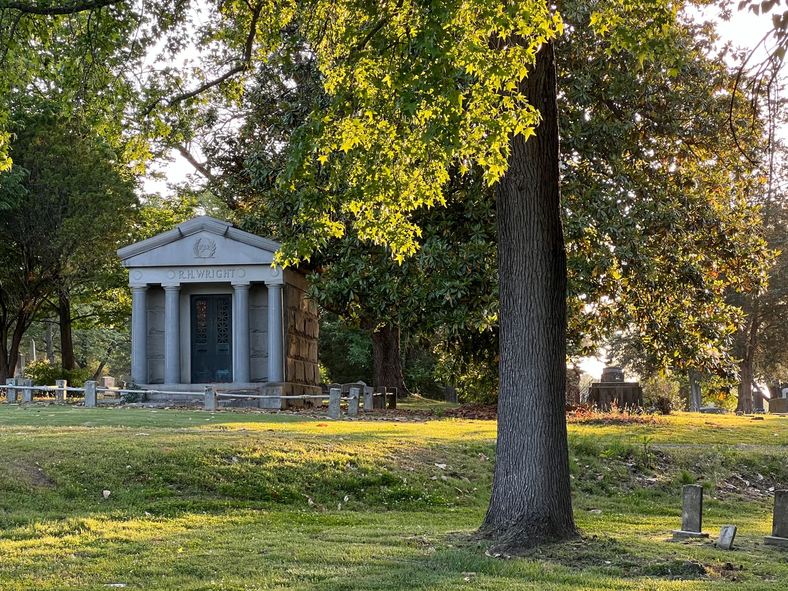 A peaceful cemetery setting features a well-kept mausoleum. The structure is surrounded by trees, with sunlight softly filtering through the leaves, illuminating the gravestones and creating long shadows on the grass.