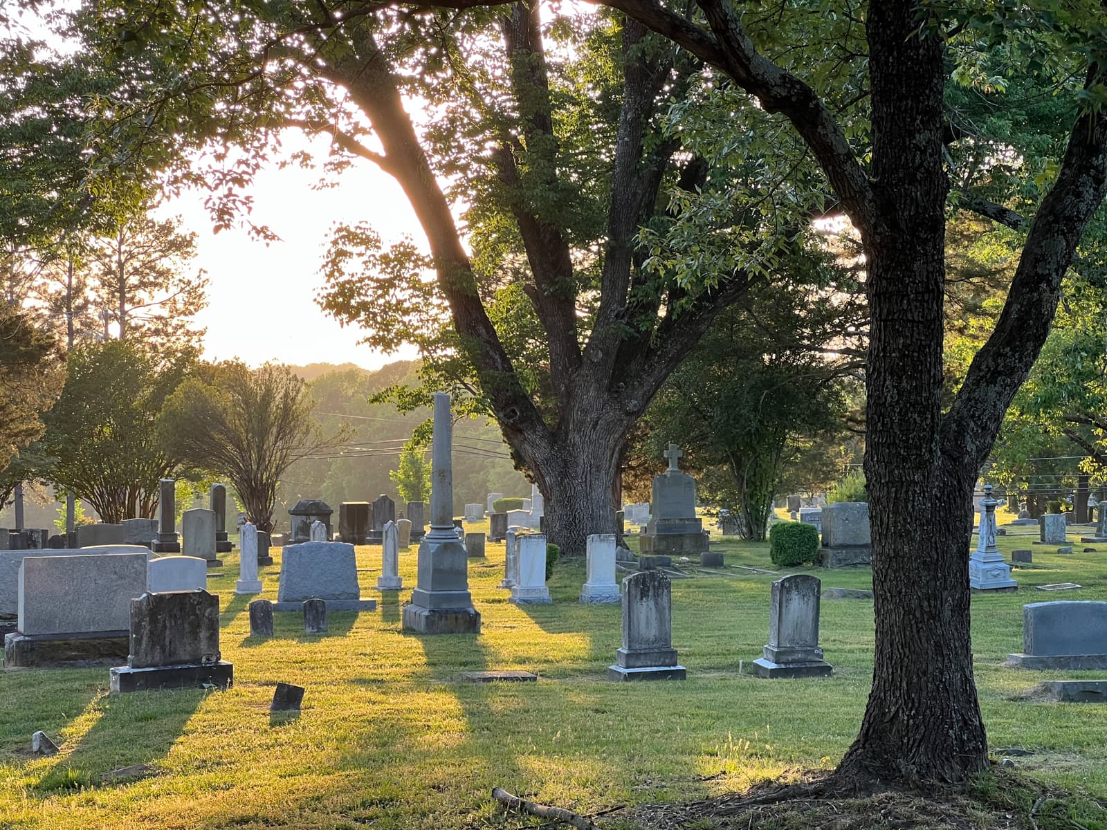 This image captures a tranquil graveyard bathed in warm, golden-hour light. The soft sunlight filters through the trees, casting long shadows across the green grass and tombstones, creating a serene and reflective atmosphere.