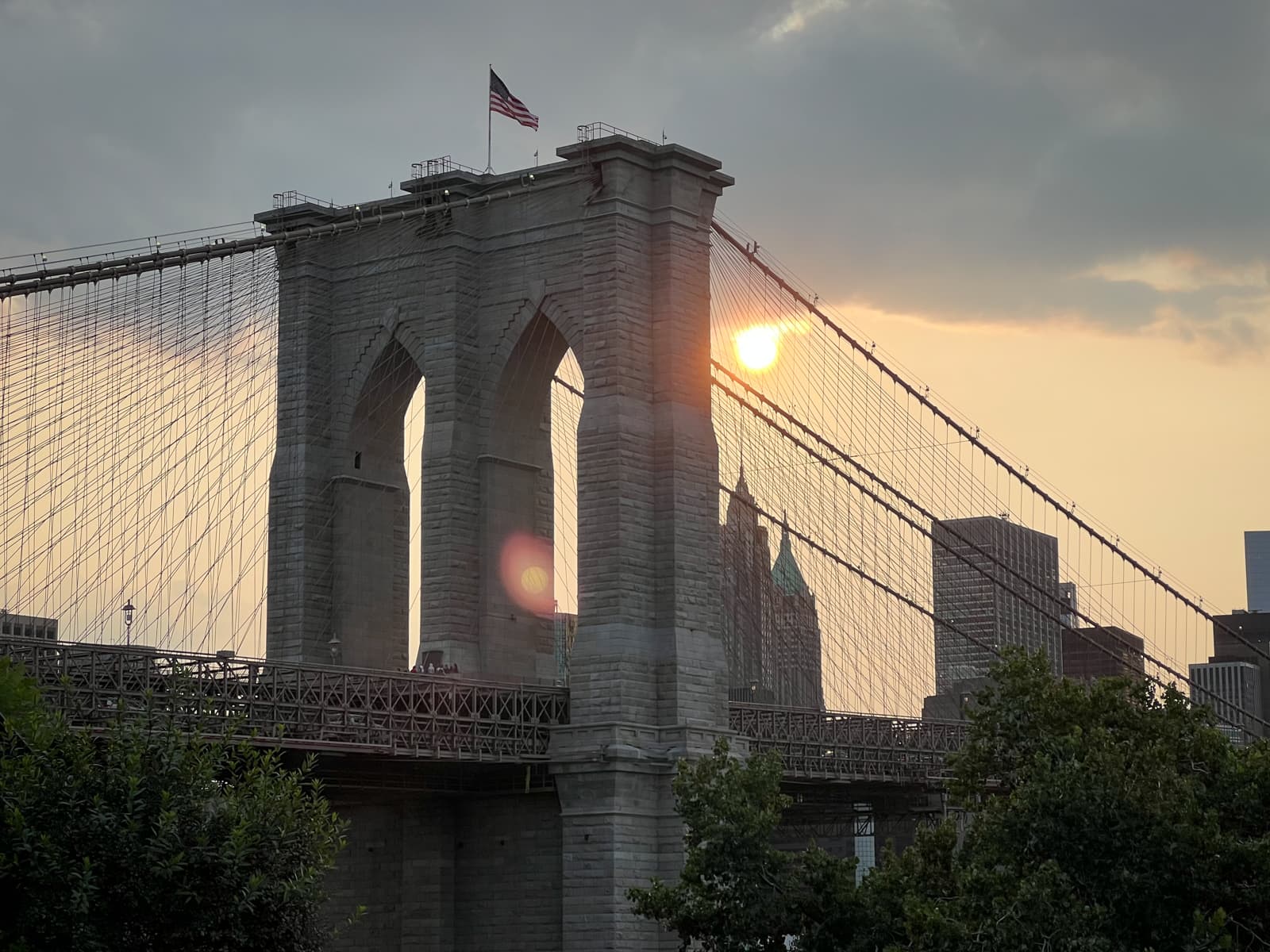 This image shows the Brooklyn Bridge, silhouetted against a setting sun. The sun peeks between the cables of the bridge, casting a warm glow over the scene. The American flag atop the bridge flutters in the breeze. In the background, the skyline of New York is visible, with some clouds overhead. The light of the setting sun and the faint lens flare create a calm, picturesque moment.
