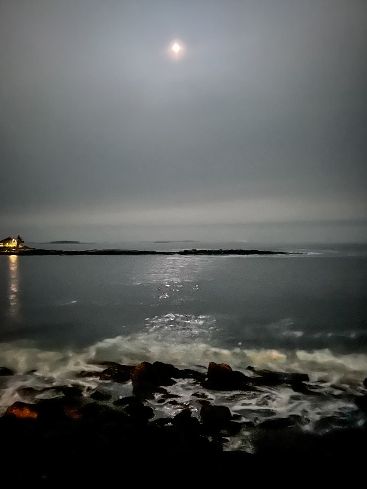 This image captures a serene nighttime scene by the sea, with the moon casting a soft, glowing light over the water. The moonlight reflects on the calm ocean, creating a shimmering pathway on the surface. In the foreground, waves crash gently against dark rocks, their frothy white tips illuminated by the moonlight. To the left, a small house or lighthouse sits near the shore, with warm lights glowing softly in contrast to the cool, muted tones of the night.