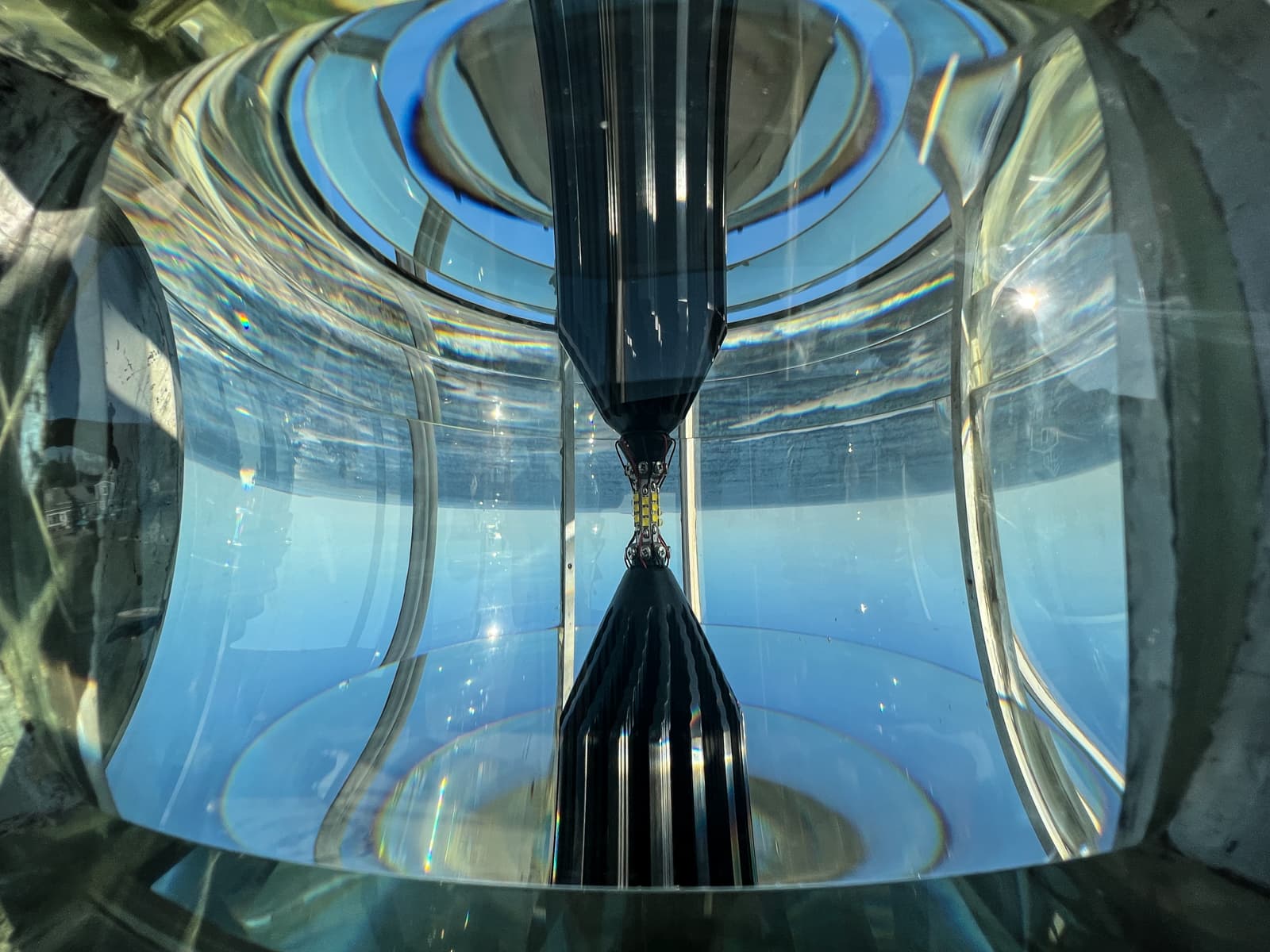 This image shows the intricate interior of a lighthouse's Fresnel lens. The reflections and refractions of light through the glass create an abstract, almost kaleidoscopic effect. The play of light and shadow, along with the curved surfaces of the lens, gives a sense of how this complex device magnifies and directs light. The view through the lens reveals a glimpse of the ocean outside, merging the technical with the natural.