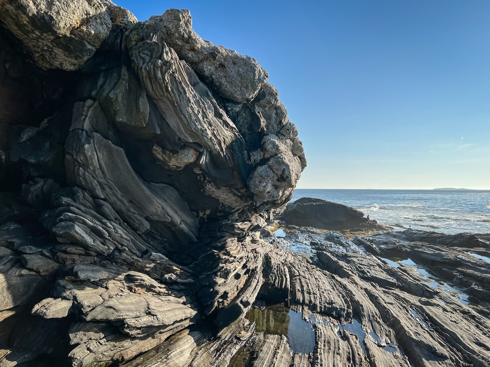 This photo captures a stunning geological formation on the coast, with intricately layered, weathered rocks. The dark striations in the rock appear almost sculptural, with the sunlight illuminating their textures. The ocean is visible in the background under a clear blue sky, adding a sense of openness and tranquility to the rugged foreground. The small pools of water trapped between the rocks reflect the light, creating an interesting contrast between the solid rock and the fluidity of the sea.
