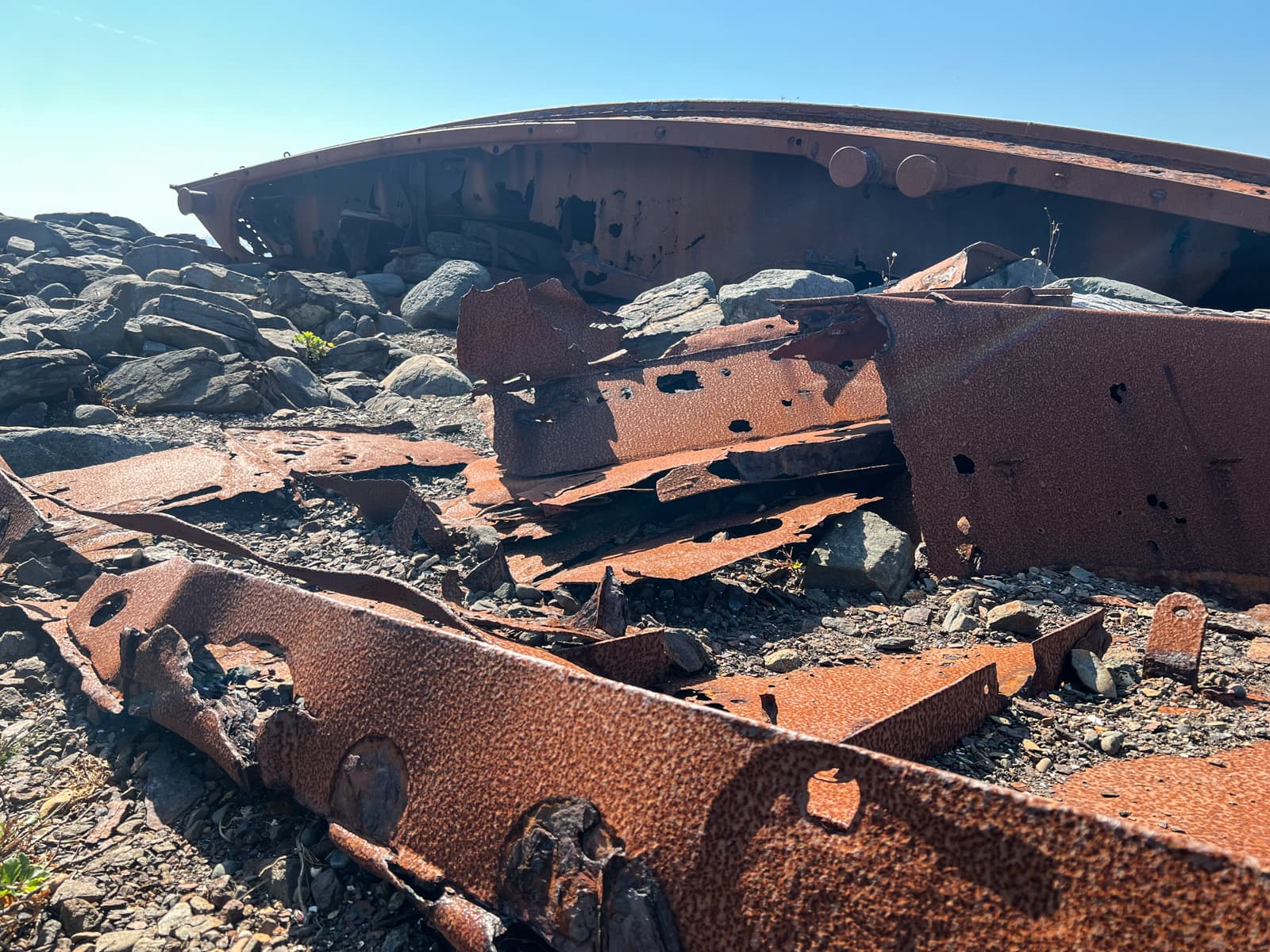 This image depicts the remains of a rusted metal structure, possibly a shipwreck or industrial debris, scattered among rocks. The decaying metal is full of holes and weathered edges, showing signs of long exposure to the elements. The bright sunlight casts shadows across the scene, enhancing the texture and the stark, broken appearance of the wreckage.