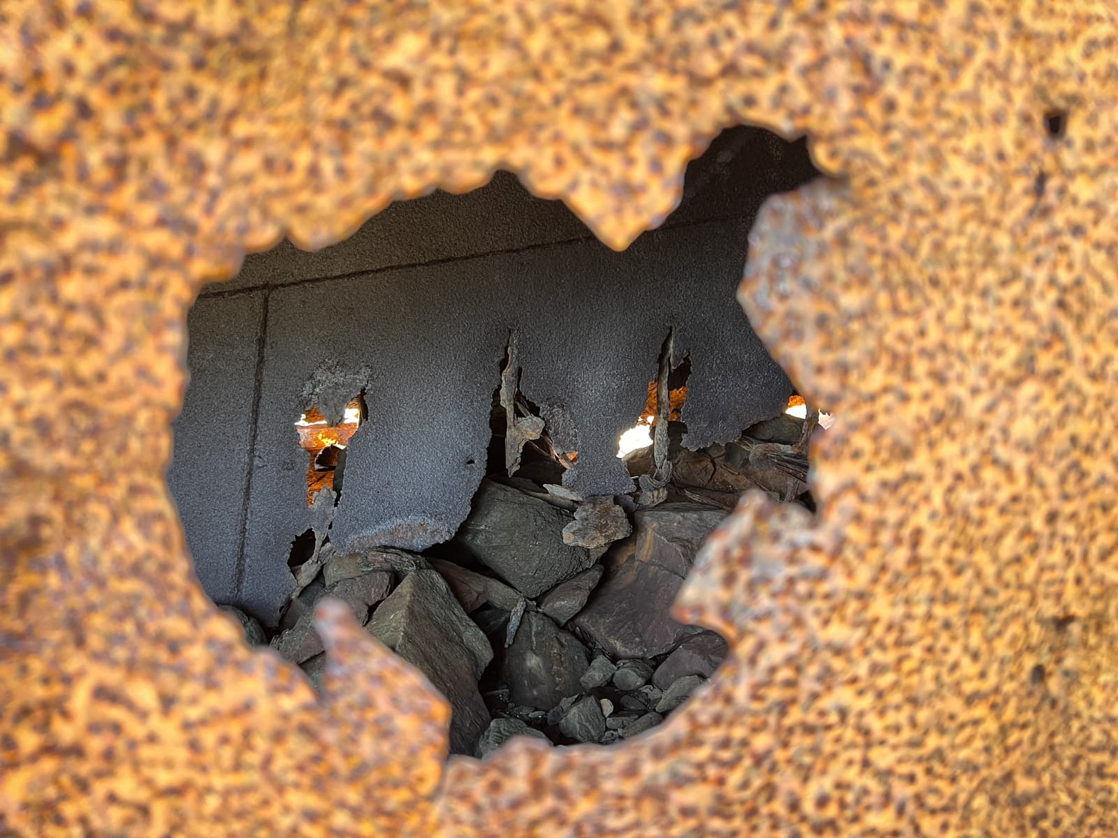 This photo captures a close-up view of a rusted metal surface with a large, jagged hole in the center. Through the hole, you can see stones and debris, creating a contrast between the rough, decaying metal and the natural elements beyond. The texture of the rusted metal gives the image a gritty, industrial feel, while the hole draws the viewer's eye to the details inside.