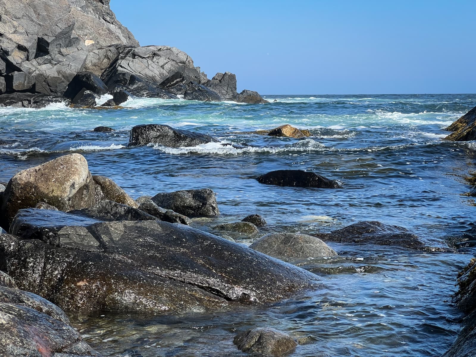 This photo shows a rugged coastal landscape with large rocks emerging from the blue-green ocean. The water is calm near the shore, with gentle waves rippling around the rocks, while larger rock formations in the distance add a sense of scale and depth. The sunlight illuminates the wet surfaces of the rocks, highlighting their textures.