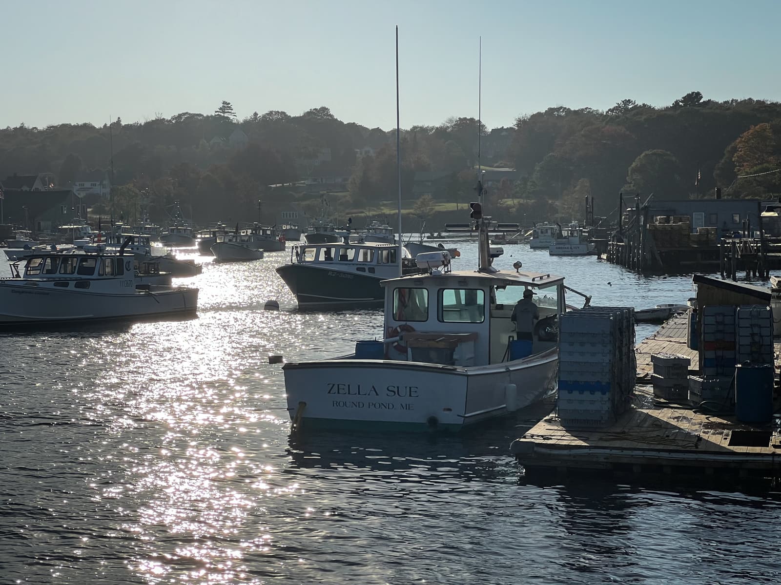 This image shows a peaceful harbor with boats gently floating on the water. The sun is low in the sky, reflecting off the water's surface and casting a warm, shimmering light. In the foreground, a boat named "Zella Sue" is docked, with stacked crates and equipment on the pier. The scene evokes a sense of quiet activity in a coastal town, with the soft morning or evening light adding to the calmness.